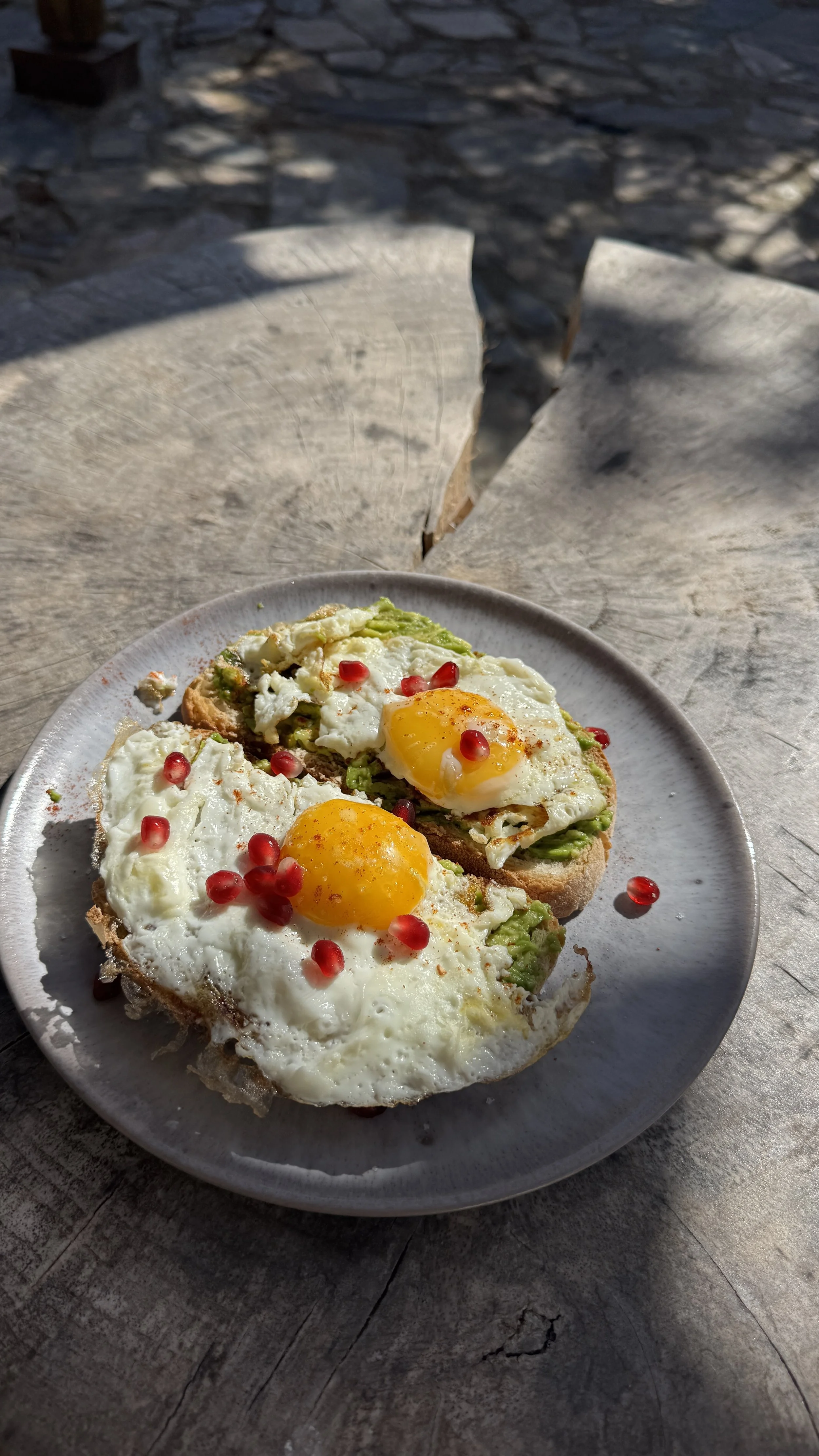 Zwei Spiegeleier auf avocadobeschmiertem Toast mit Granatapfelkernen, auf einem weißen Teller, im Freien bei Sonnenschein auf einem Holztisch