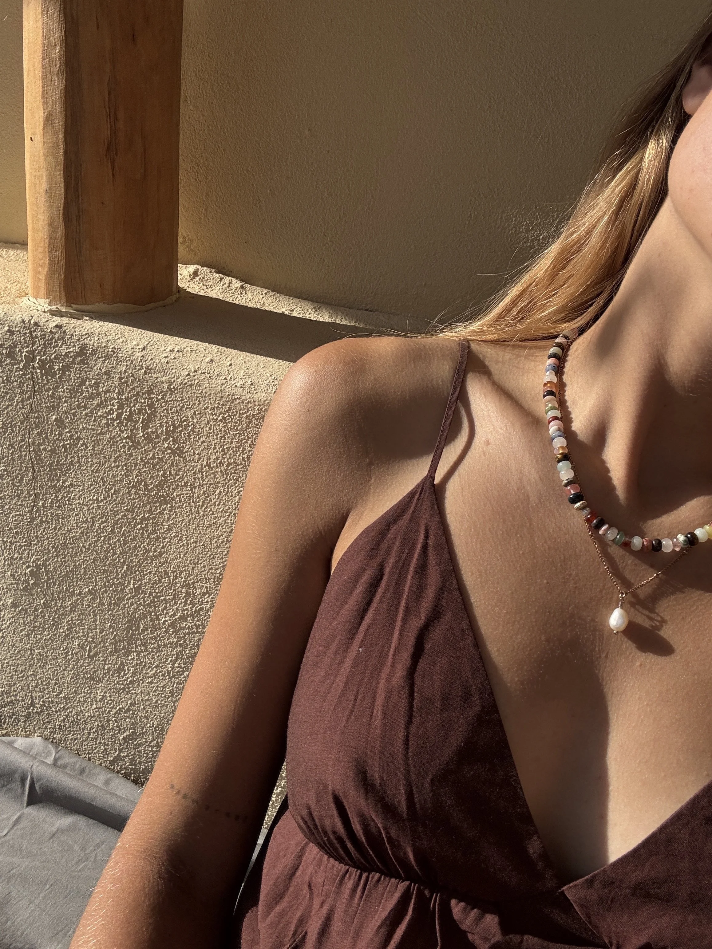 Nahaufnahme eines Frauenoberkörpers, die Schulter, ein Teil des Gesichts und die Halskette mit Perlen und Anhänger sind sichtbar, Sonnenlicht fällt auf die Haut, Hintergrund mit einer Wand und Holzrahmen.