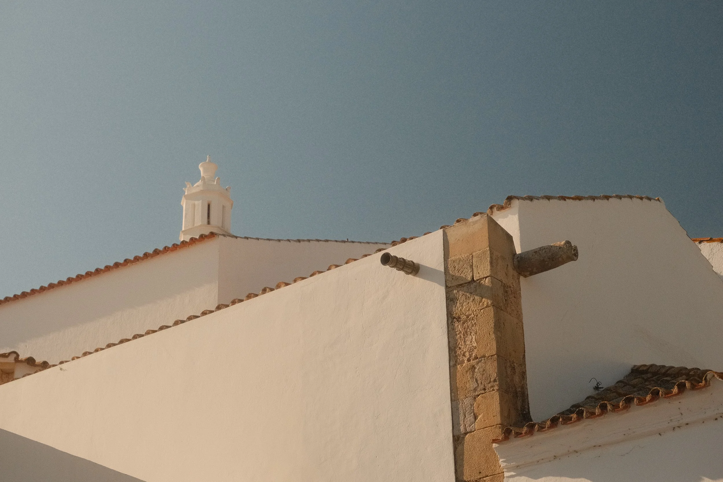 Weißes Gebäude mit Ziegeldach und Kirchturm im Hintergrund, blauer Himmel.