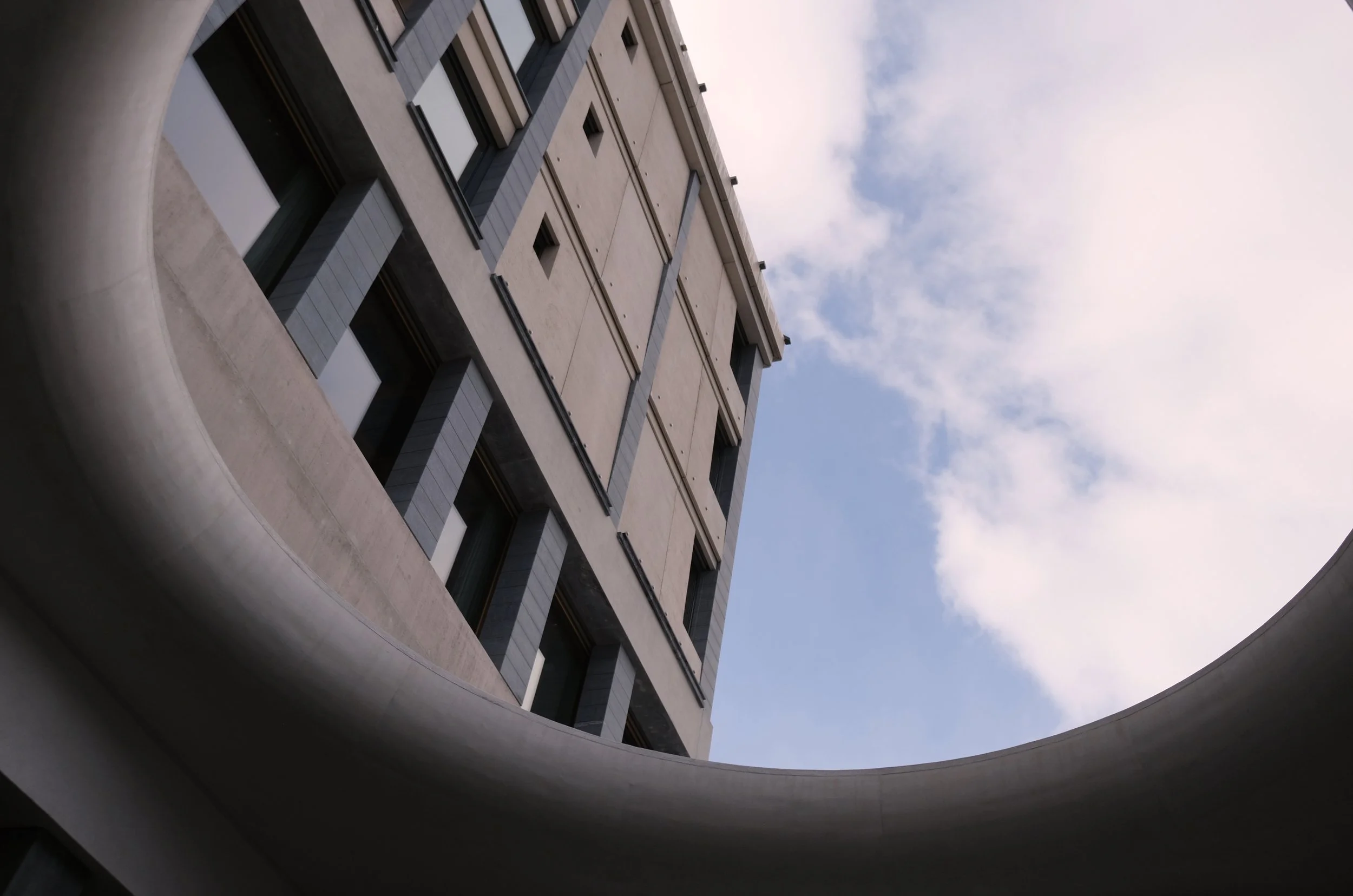 Ansicht eines modernen Gebäudes von unten, umgeben von einem runden, hellgrauen Bauwerk, mit Blick nach oben zu blauen Himmel mit Wolken.