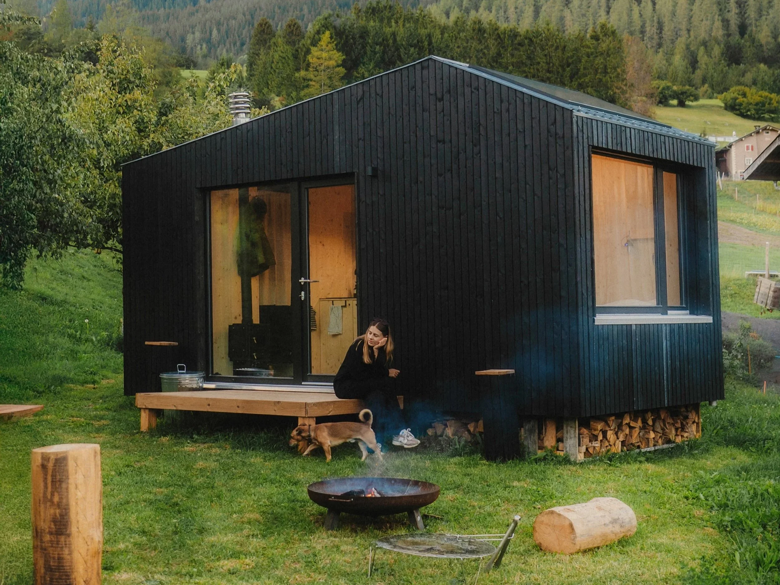 Ein modernes schwarzes Holzhaus mit großen Fenstern in einer grünen Landschaft. Im Vordergrund sitzt eine junge Frau neben einem kleinen Hund, es gibt einen Feuerkorb und Holzstämme im Garten.
