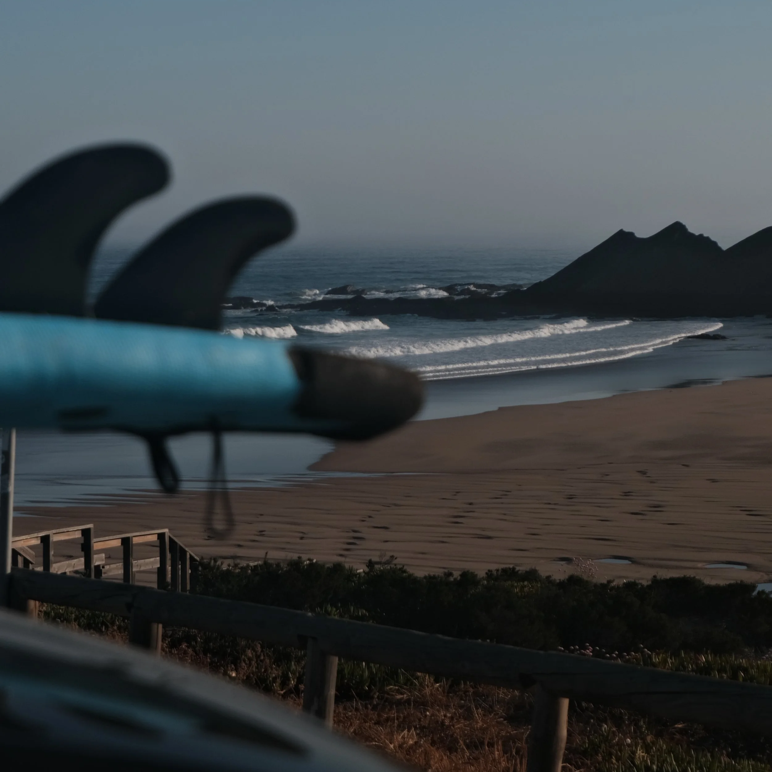 Ein Strand mit Wellen, Felsen im Hintergrund, und eine Surfbretthalterung im Vordergrund.