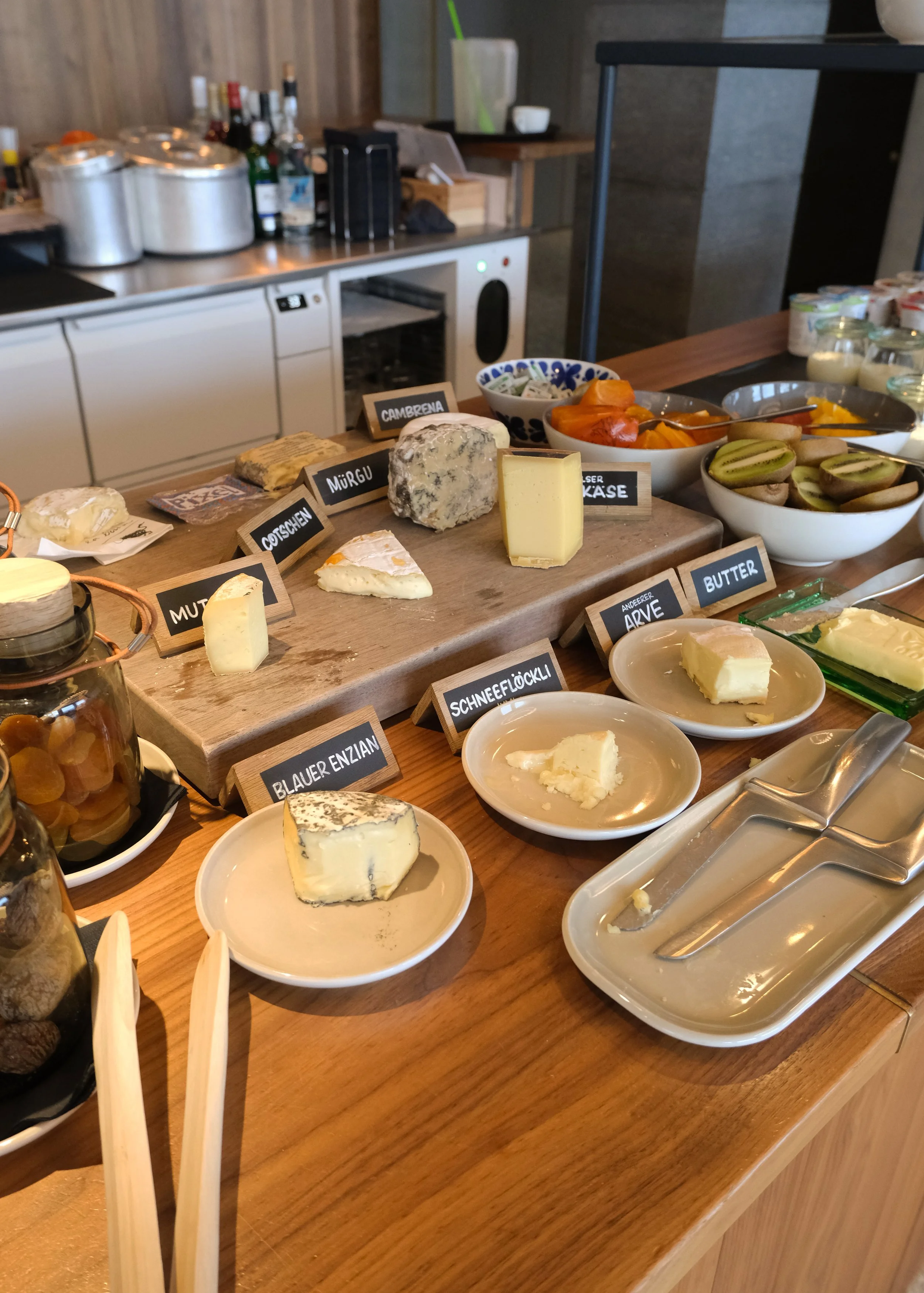 Verschiedene Käsesorten auf Tafel und Tellern, darunter Blauschimmelkäse, Camembert, und andere, mit Bezeichnungen auf kleinen Schildern, im Hintergrund warme Speisen und frisches Obst in Schalen.