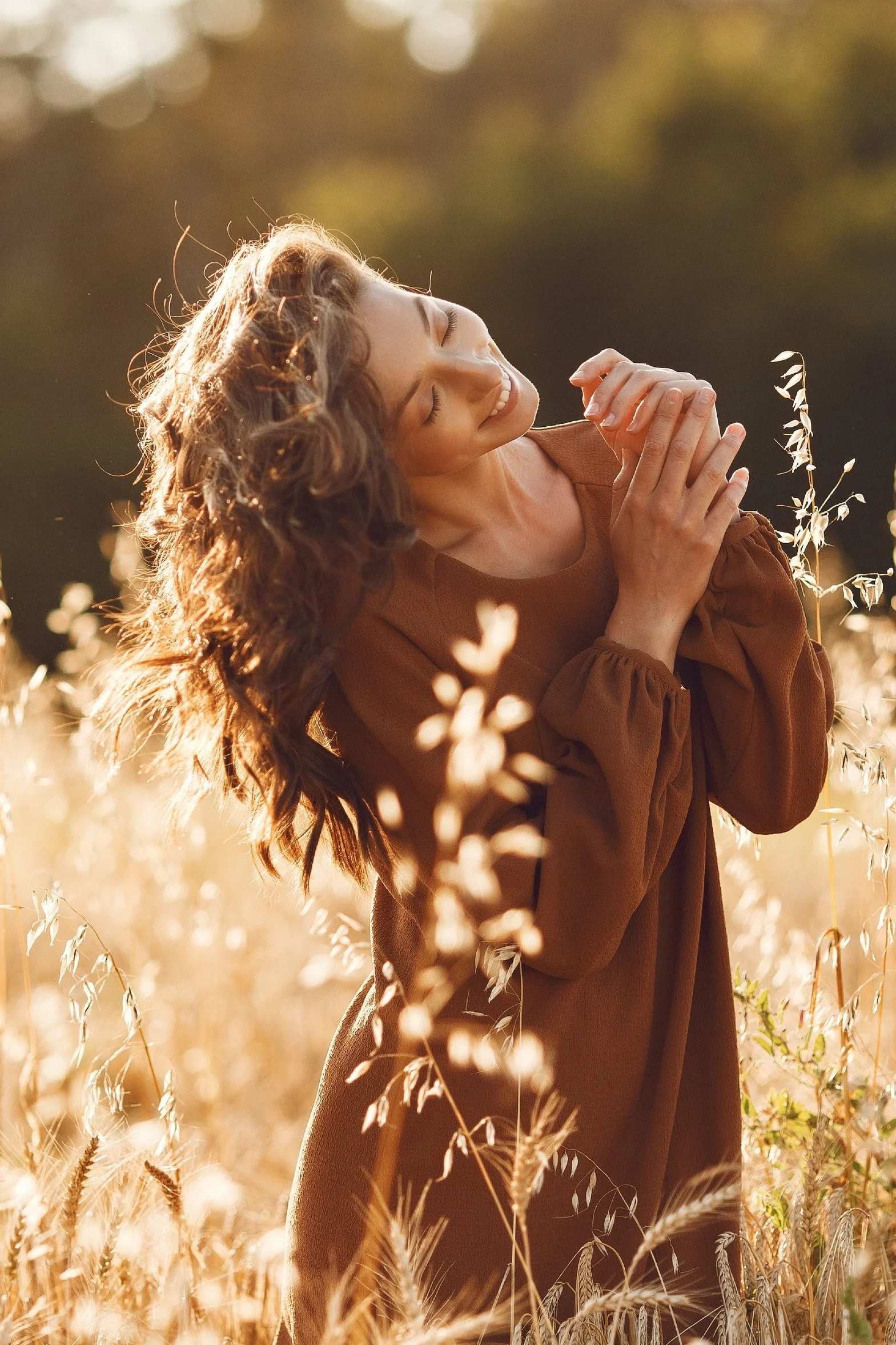 Frau in braunem Kleid in einem Weizenfeld bei Sonnenuntergang, lächelnd und mit der Hand im Haar.
