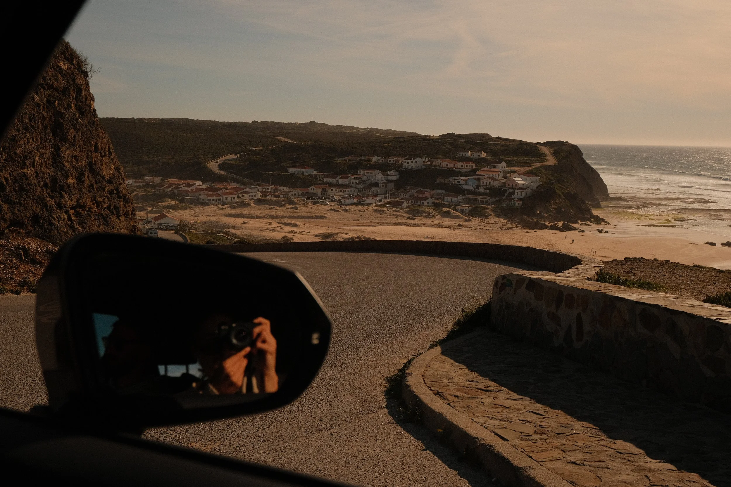Blick aus einem Auto auf eine Küstenstraße mit Blick auf eine kleine Siedlung am Strand und das Meer bei Sonnenuntergang.