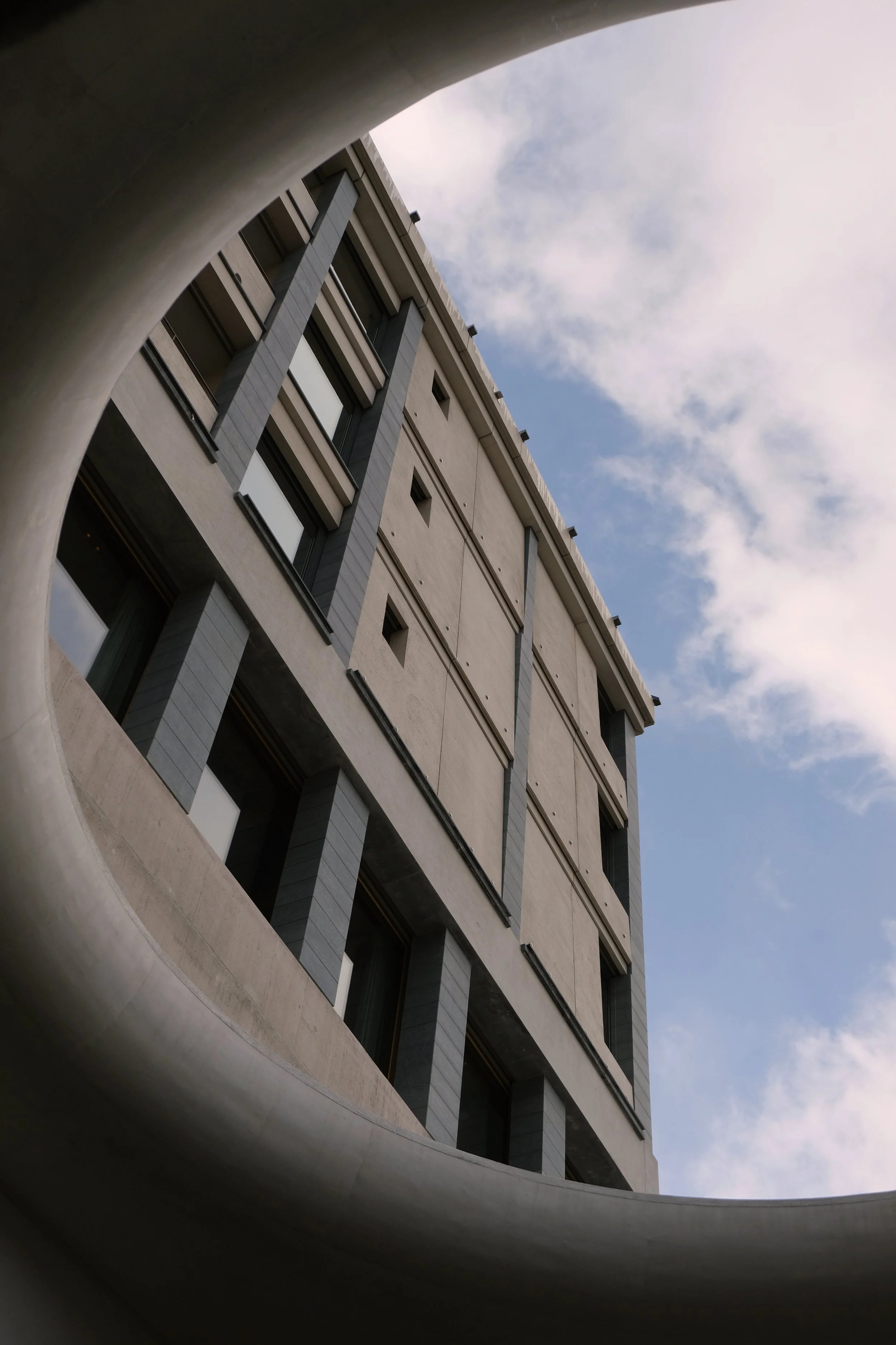 Aufnahme eines modernen Hochhauses, fotografiert von unten durch eine runde Öffnung im Vordergrund, mit Himmel und Wolken im Hintergrund.