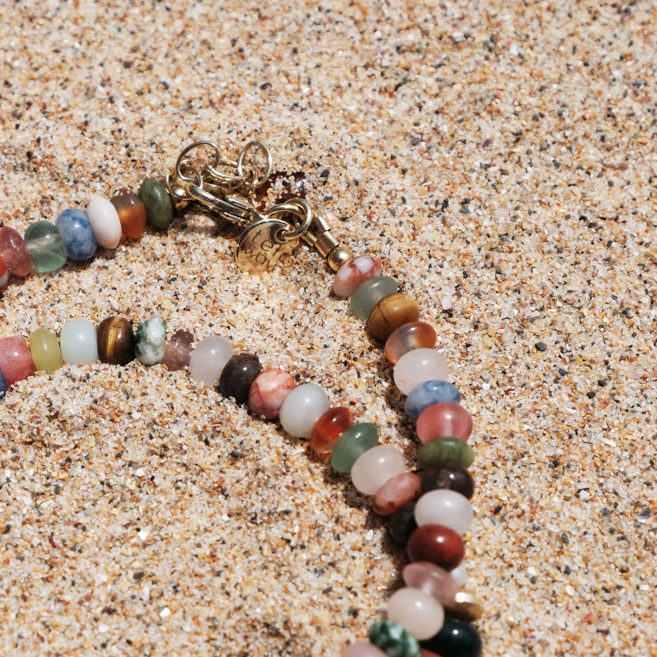 Eine bunte Perlenkette liegt im Sand, die mit einem goldfarbenen Verschluss versehen ist.