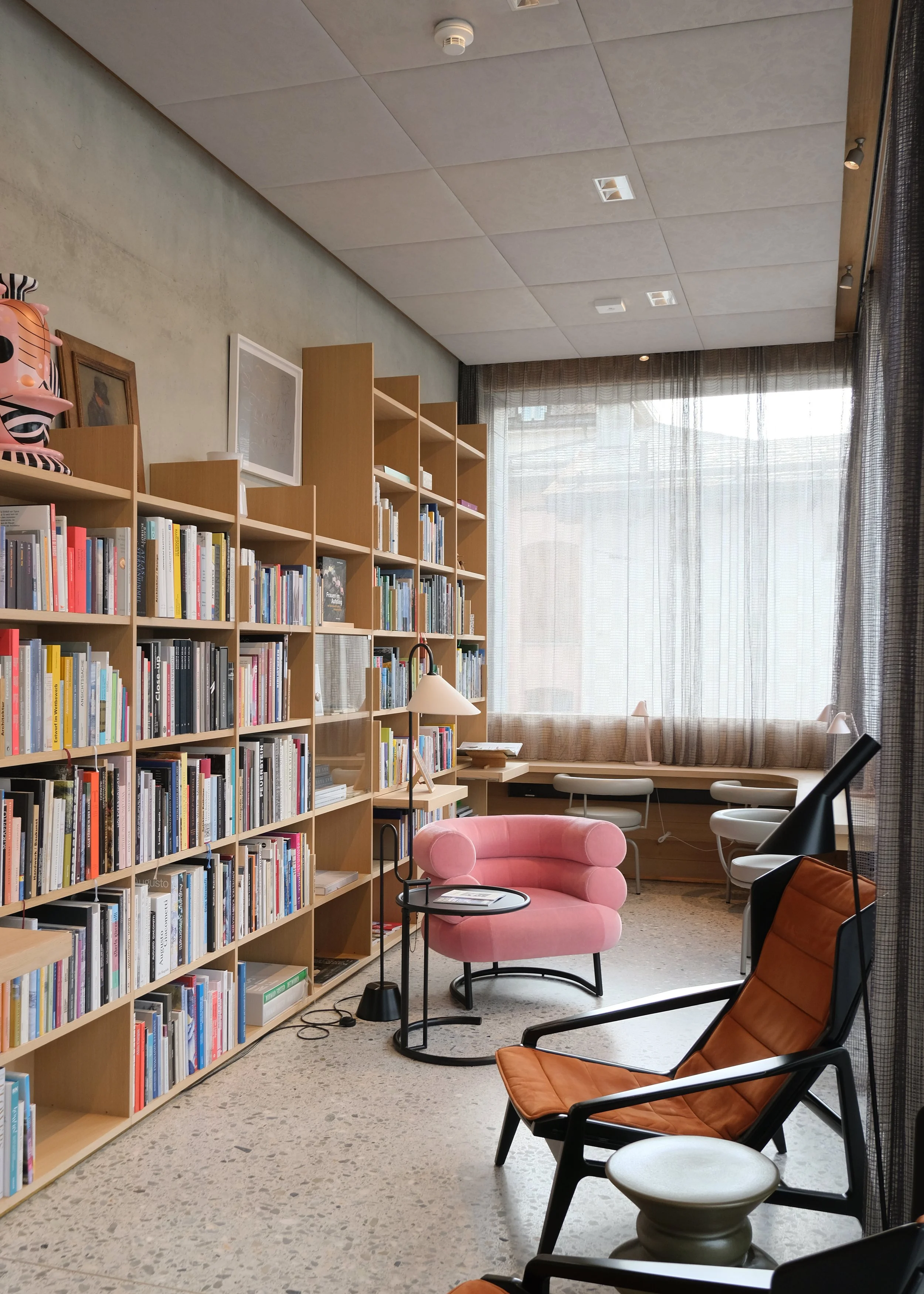 Gemütliches Lesezimmer mit Bücherregalen, Sessel, einem pinken Sofa, einem kleinen Beistelltisch, Stehlampen und einem großen Fenster mit Vorhängen.