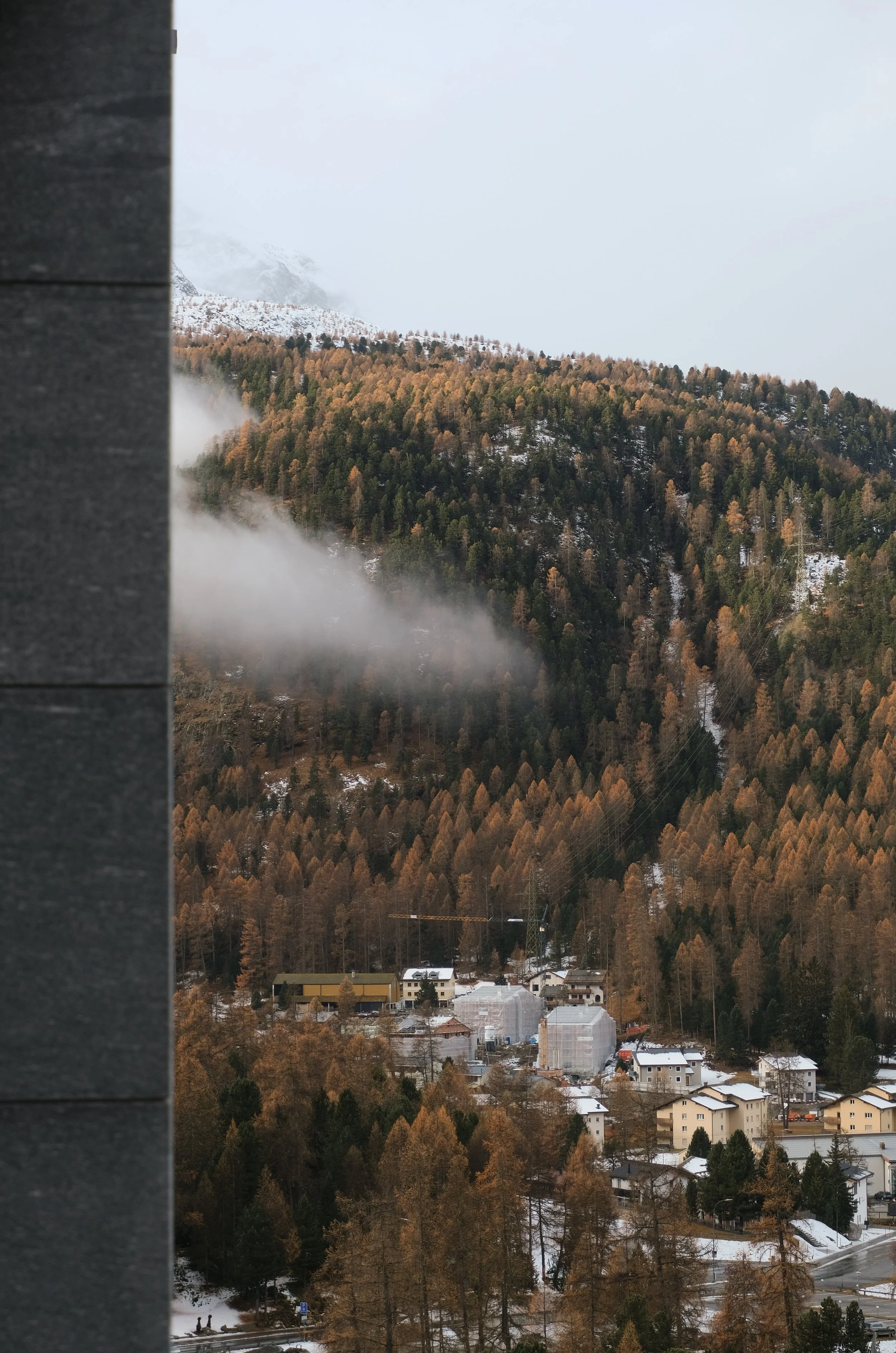 Bergdorf in den Bergen mit verschneiten Bäumen, Nebel und Wolken am Himmel, einige Häuser im Vordergrund.
