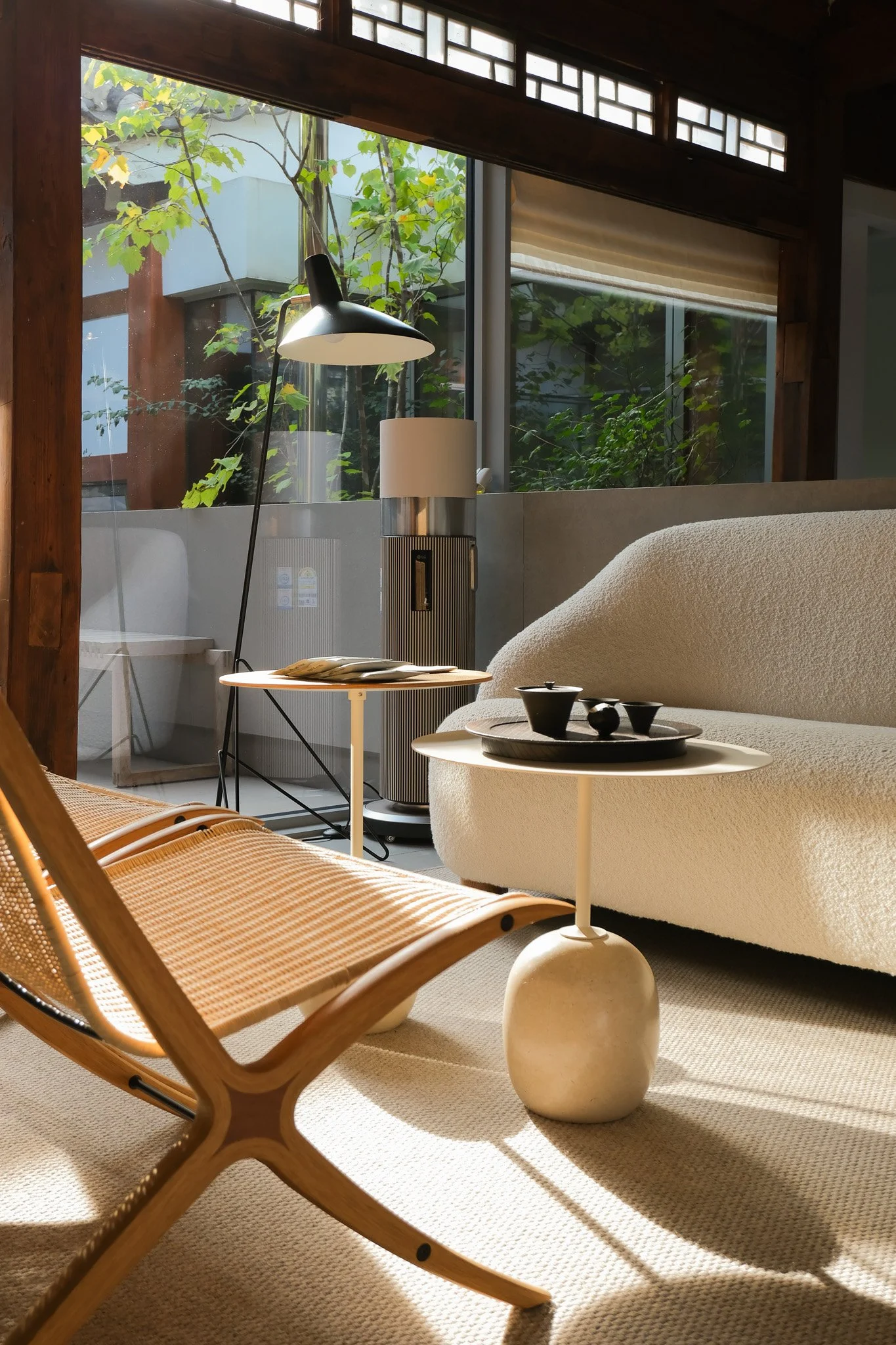Ein gemütliches Wohnzimmer mit großen Fenstern, einem weißen Sofa, einem kleinen runden Beistelltisch mit schwarzen Keramikschalen, einem modernen Lampenschirm, einem Pflanzenbild im Hintergrund und natürlichem Licht.