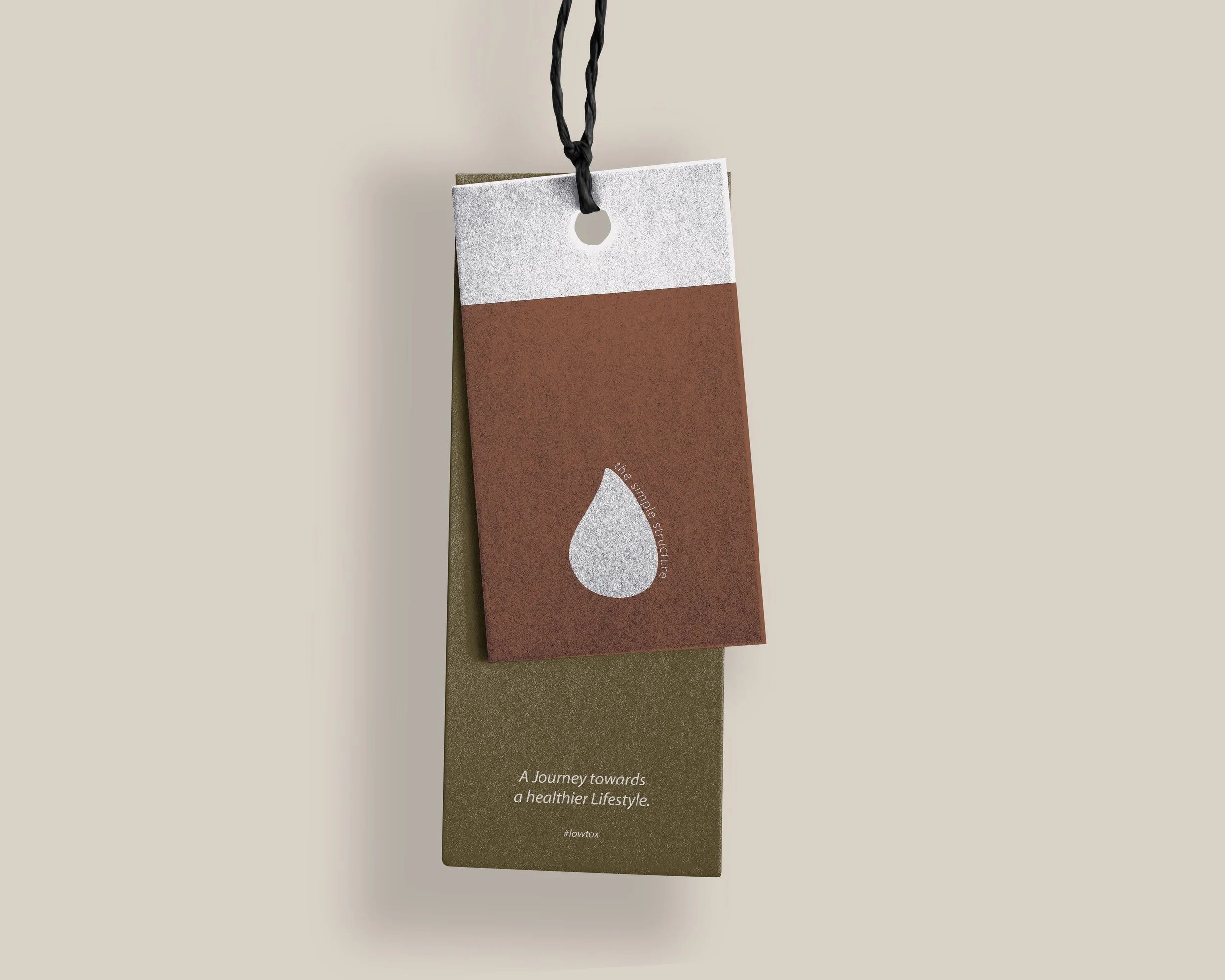 Zwei an einer Schnur befestigte Etiketten übereinander. Das obere Etikett ist braun mit einem Tropfen-Symbol, das untere grün mit dem Text "A Journey towards a healthier Lifestyle. #lowtox".
