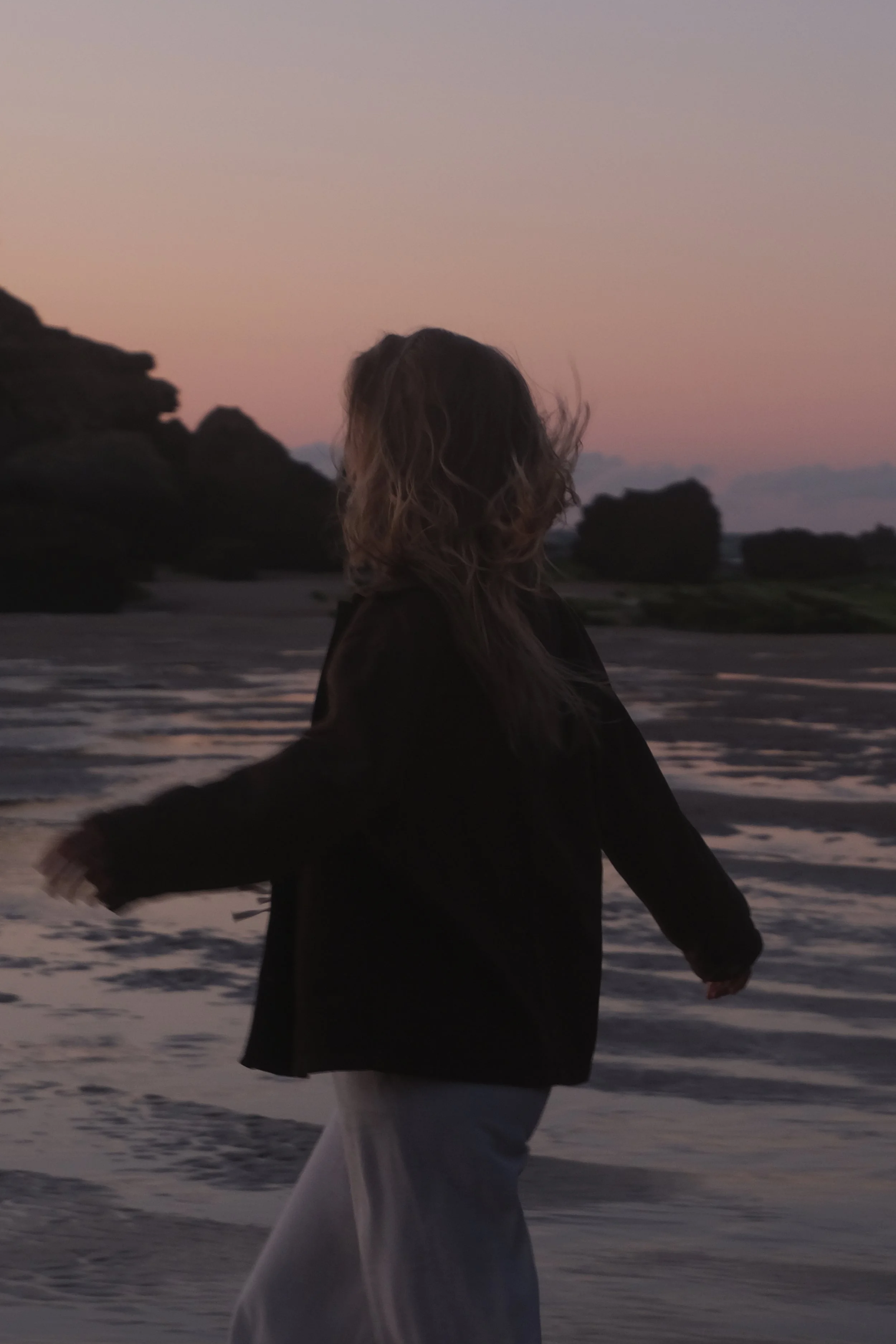 Eine Person steht am Strand bei Sonnenuntergang und blickt aufs Meer, trägt dunkle Jacke und helle Hose, mit Wind im Haar.