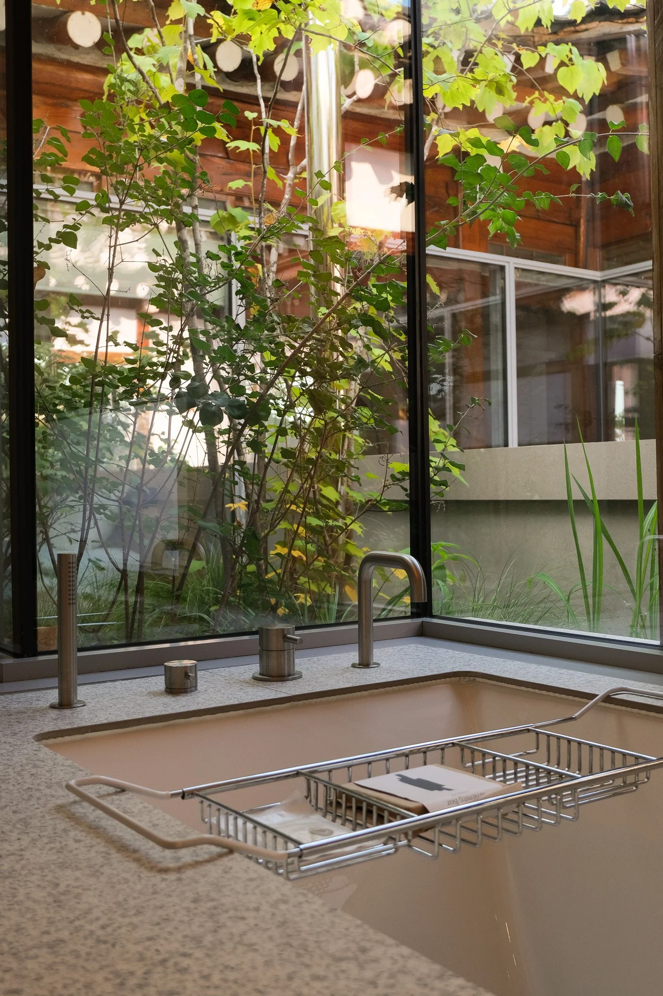 Innenaufnahme einer Küche mit einem Wasserhahn und einem Edelstahl-Geschirrkorb auf der Arbeitsplatte, Blick durch ein großes Fenster auf einen Garten mit Bäumen.