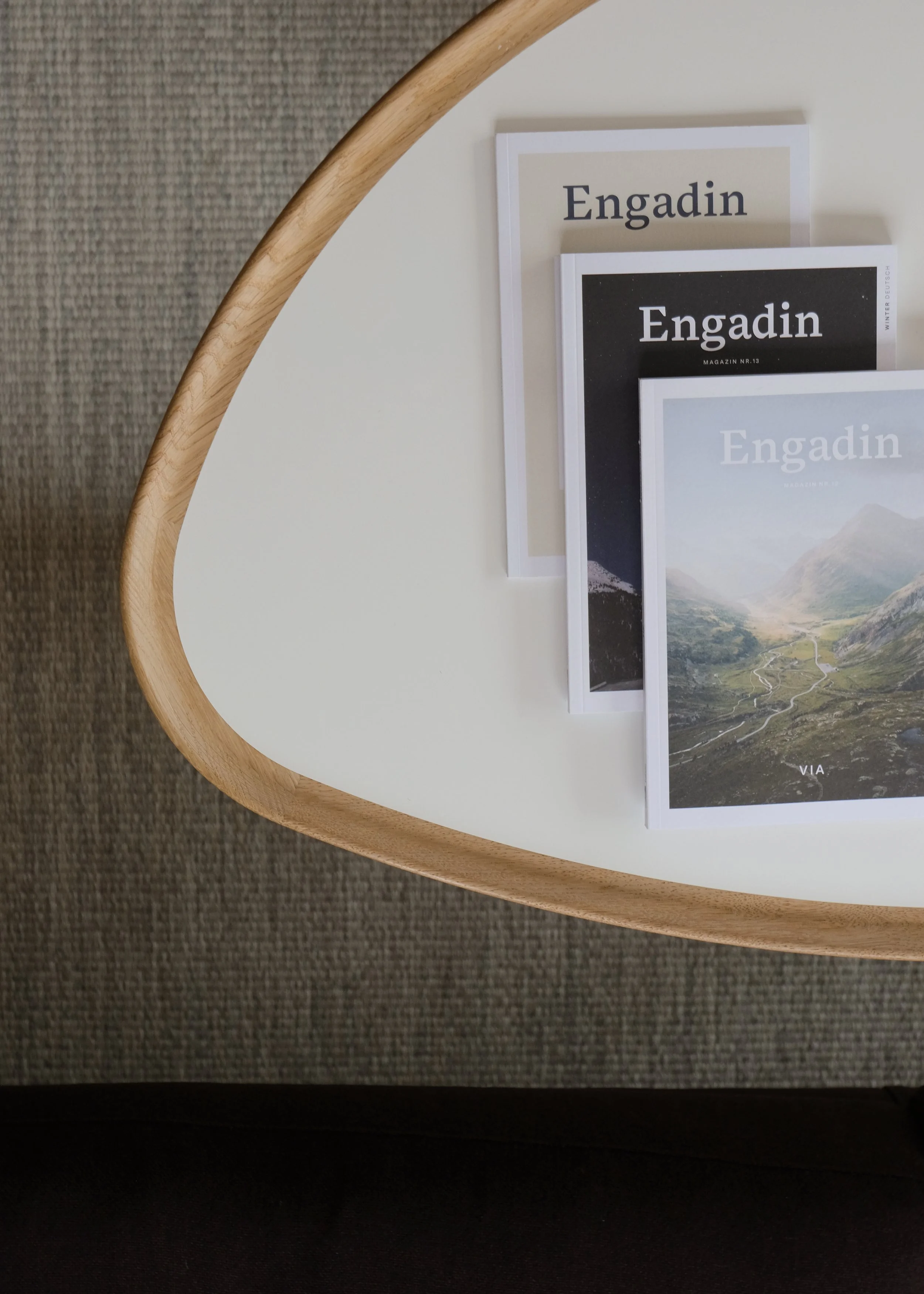 Drei Magazine mit dem Titel "Engadin" liegen auf einem weißen Tisch mit einer Holzrahmen. Das Magazin zeigt Landschaftsbilder aus den Bergen.