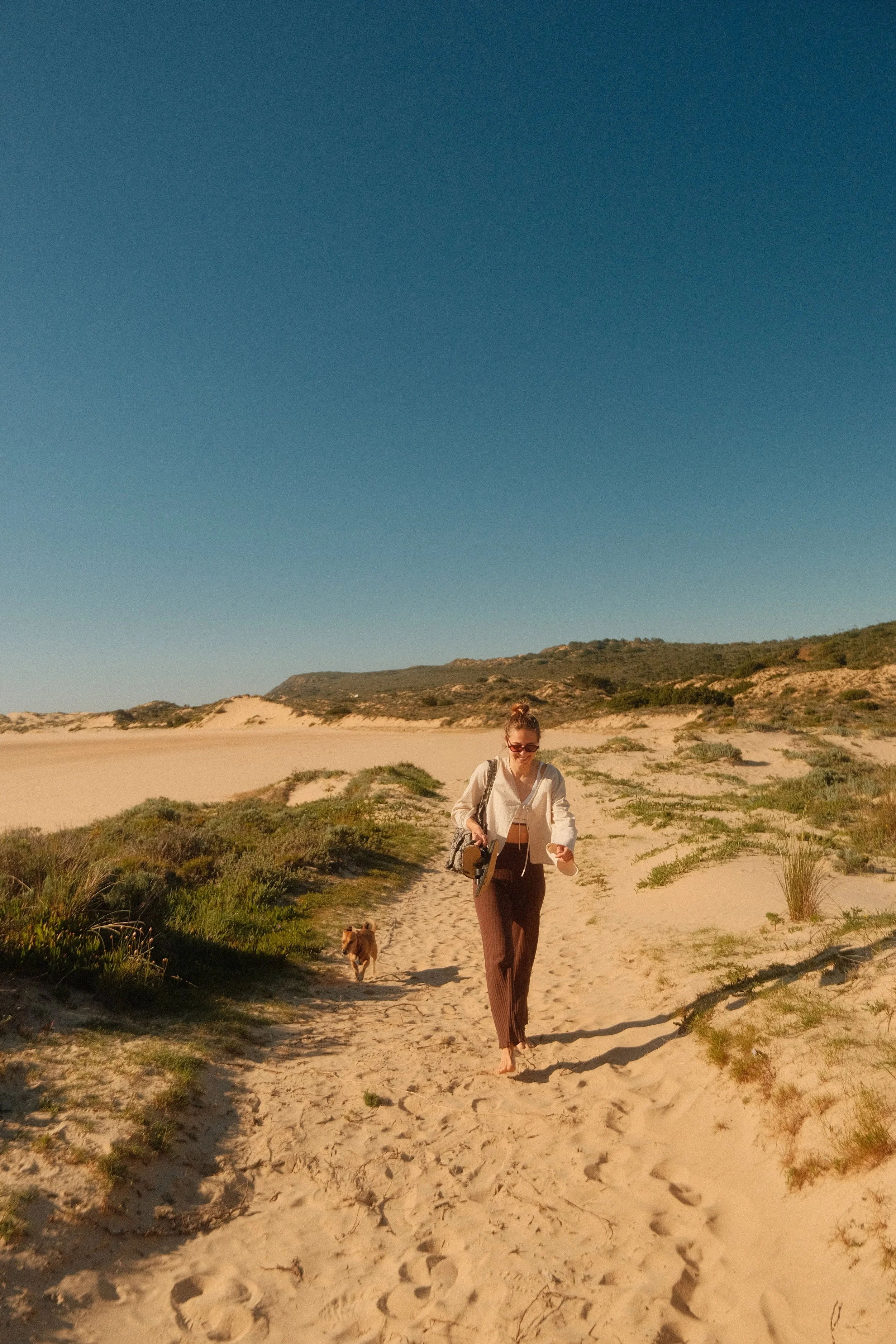 Eine Frau geht barfuß am Strand entlang, begleitet von einem kleinen Hund. Im Hintergrund sind Sanddünen und grüne Pflanzen bei hellem Sonnenschein zu sehen.