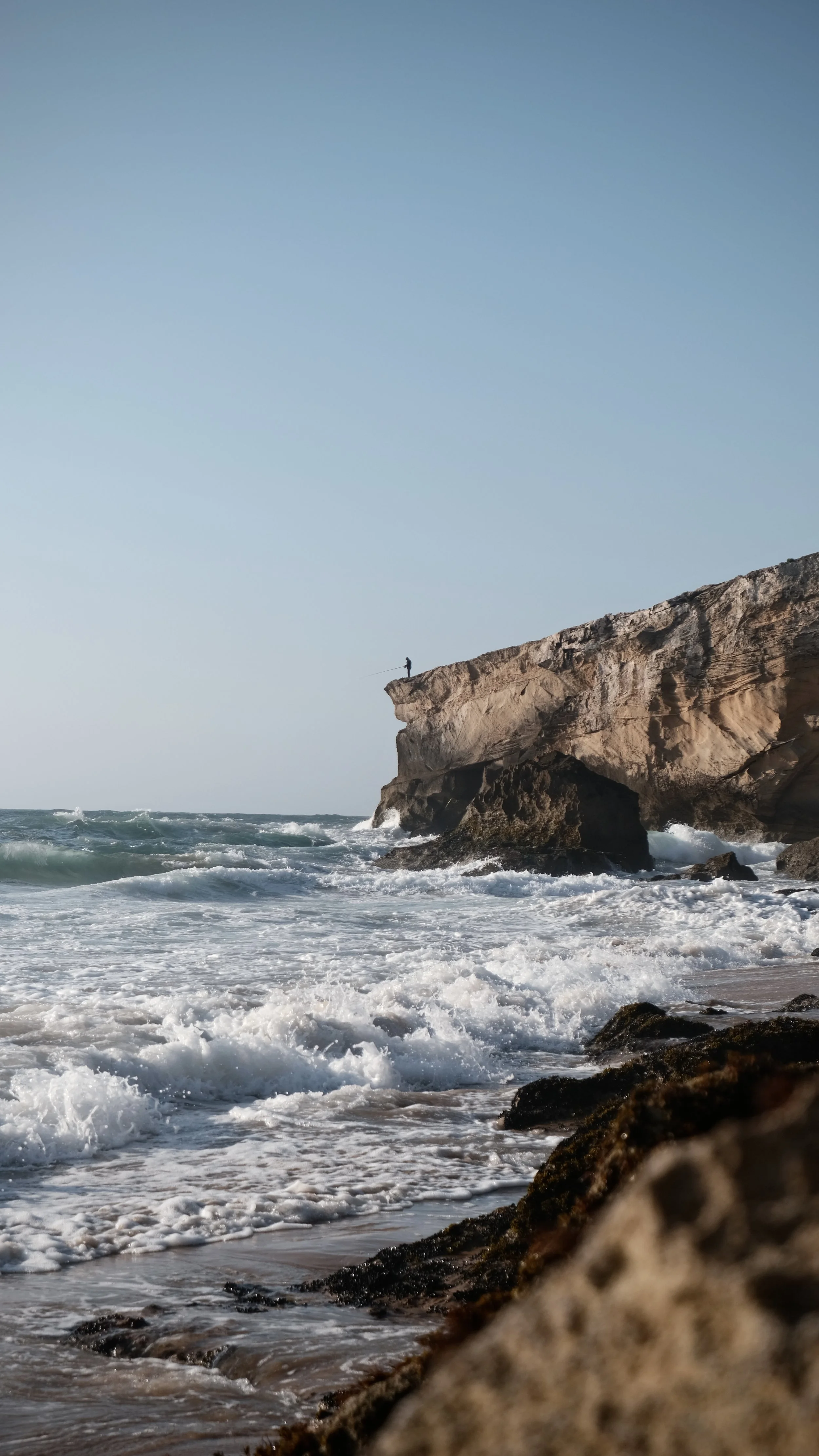Ein einzelner Mensch steht auf einer Klippe am Strand, Blick auf das Meer mit Wellen, Felsen im Vordergrund, blauer Himmel.