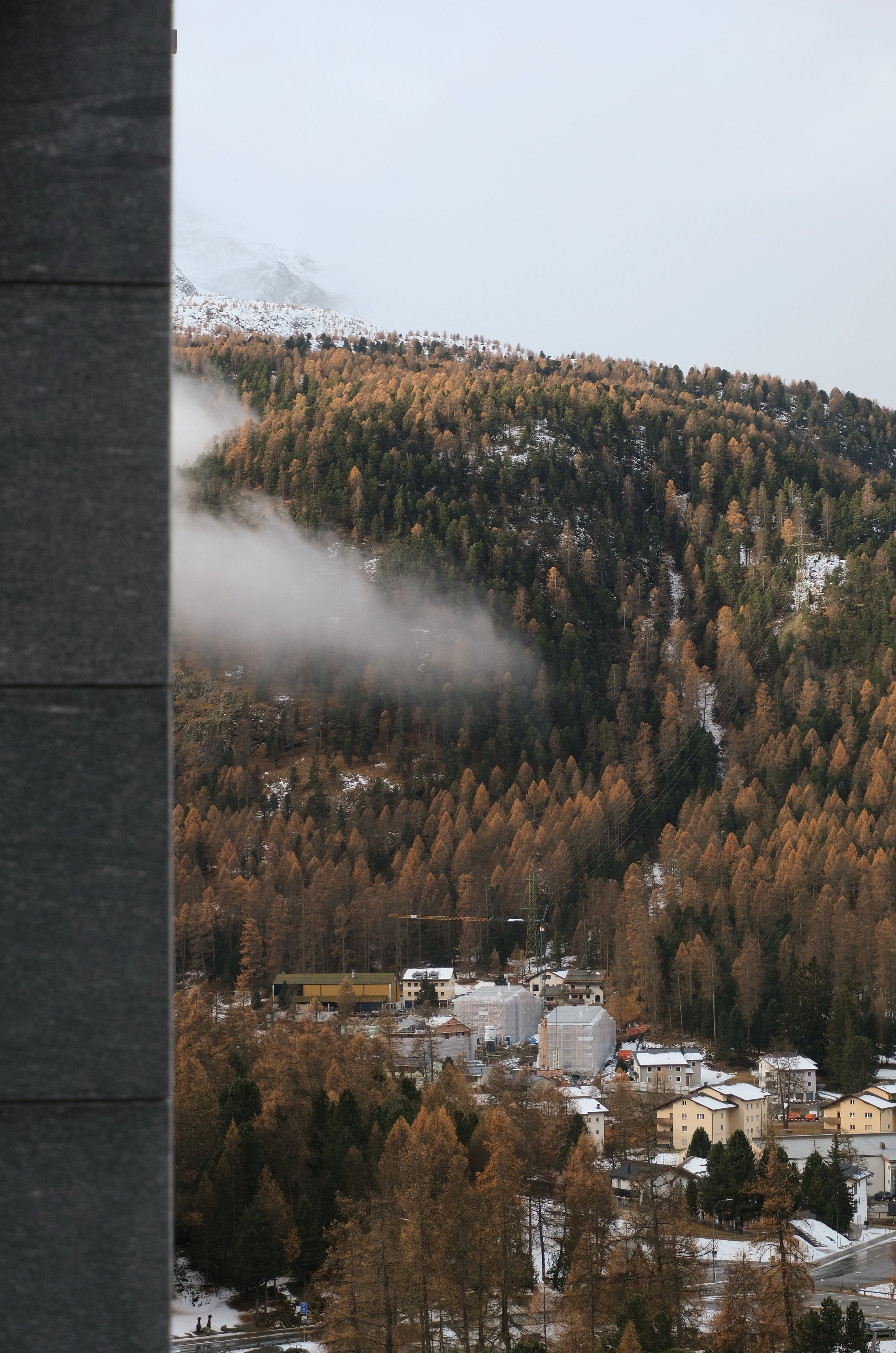 Blick auf eine verschneite Hügellandschaft mit Bäumen in Herbstfarben, kleine Häuser im Tal und Nebel um die Berge.