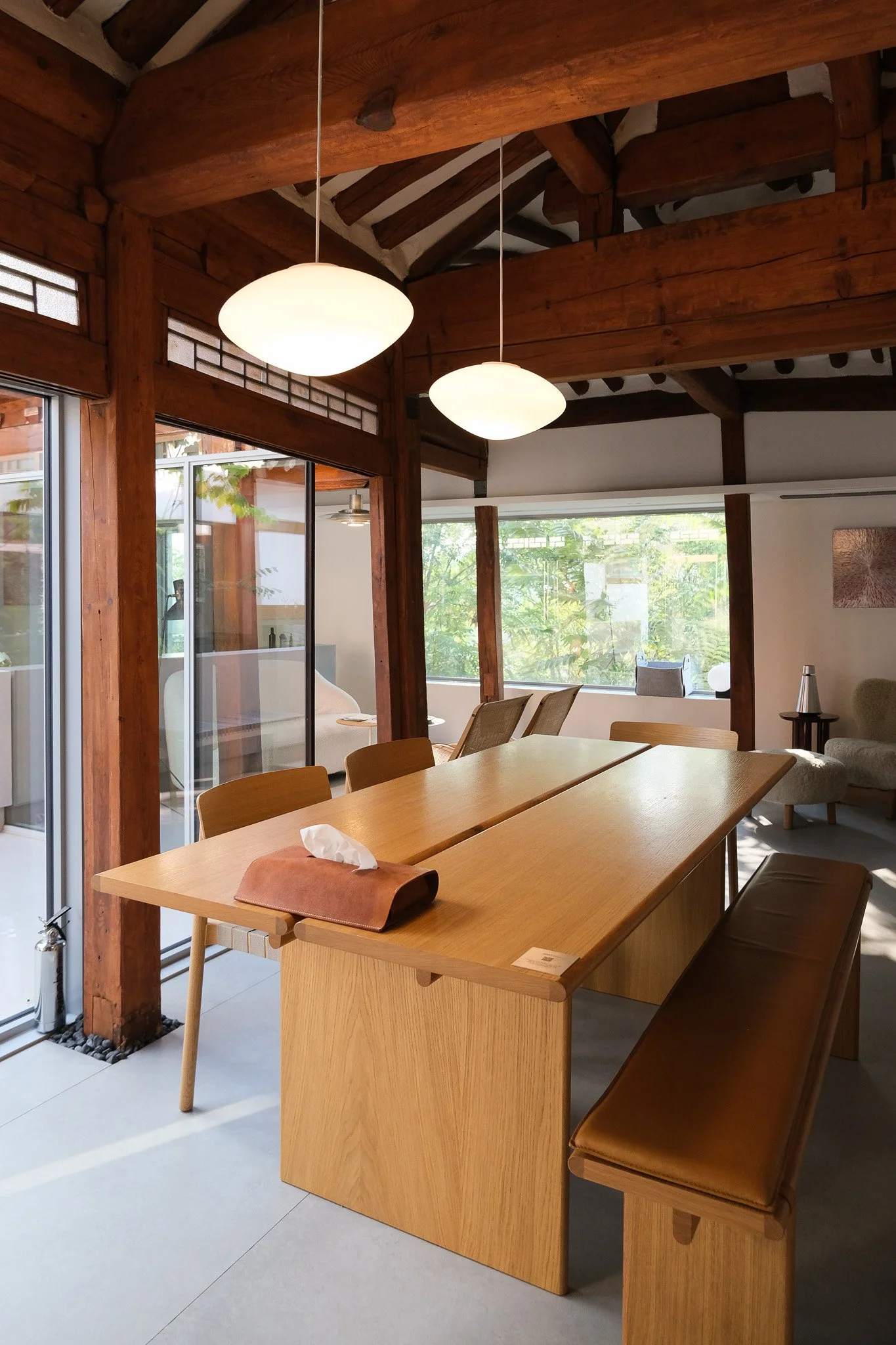 Innenraum eines Raums mit Holzdecke, Holzwänden und einem großen Holztisch, der von Stühlen umgeben ist, natürlichem Licht durch große Fenster und modernen Lampen an der Decke.