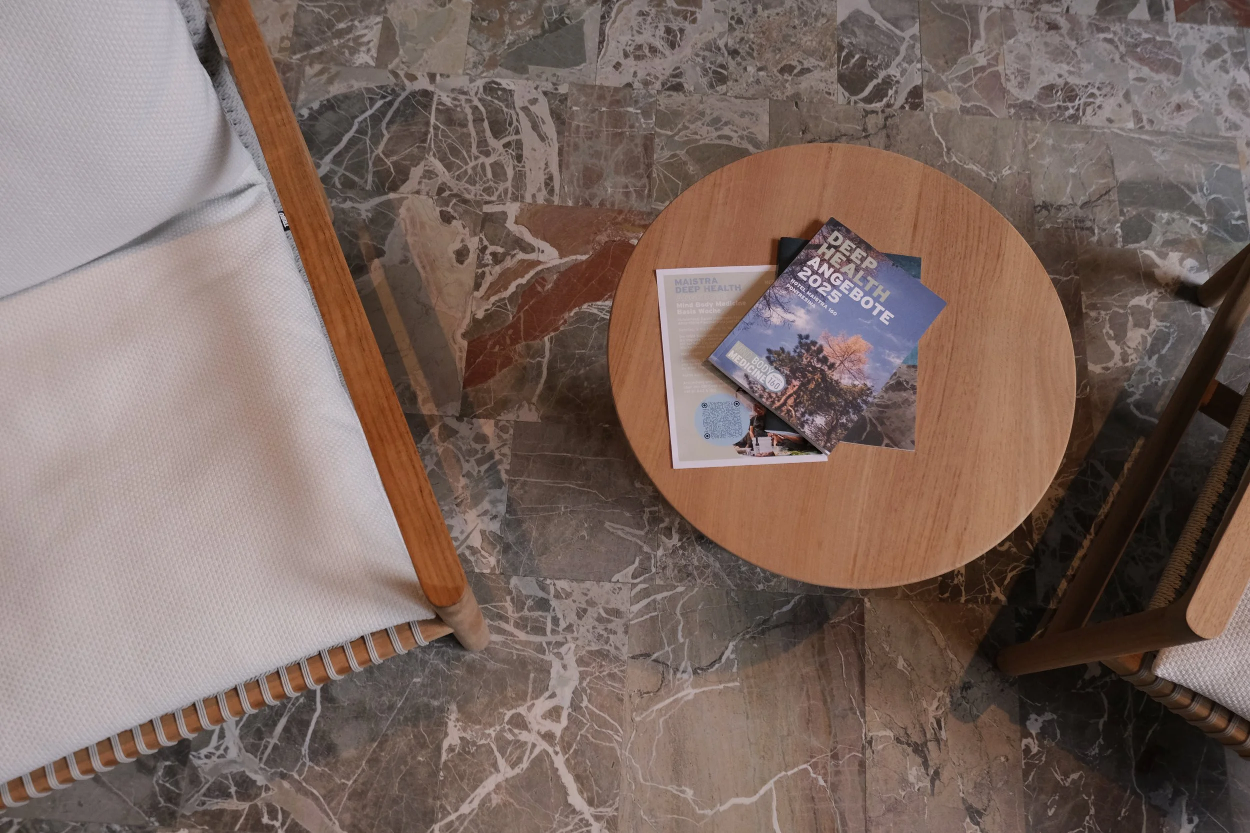 Ein runder Holztisch mit zwei Broschüren und einer Zeitschrift darauf, umgeben von zwei Stühlen mit weißen Kissen auf einem Marmorfußboden.