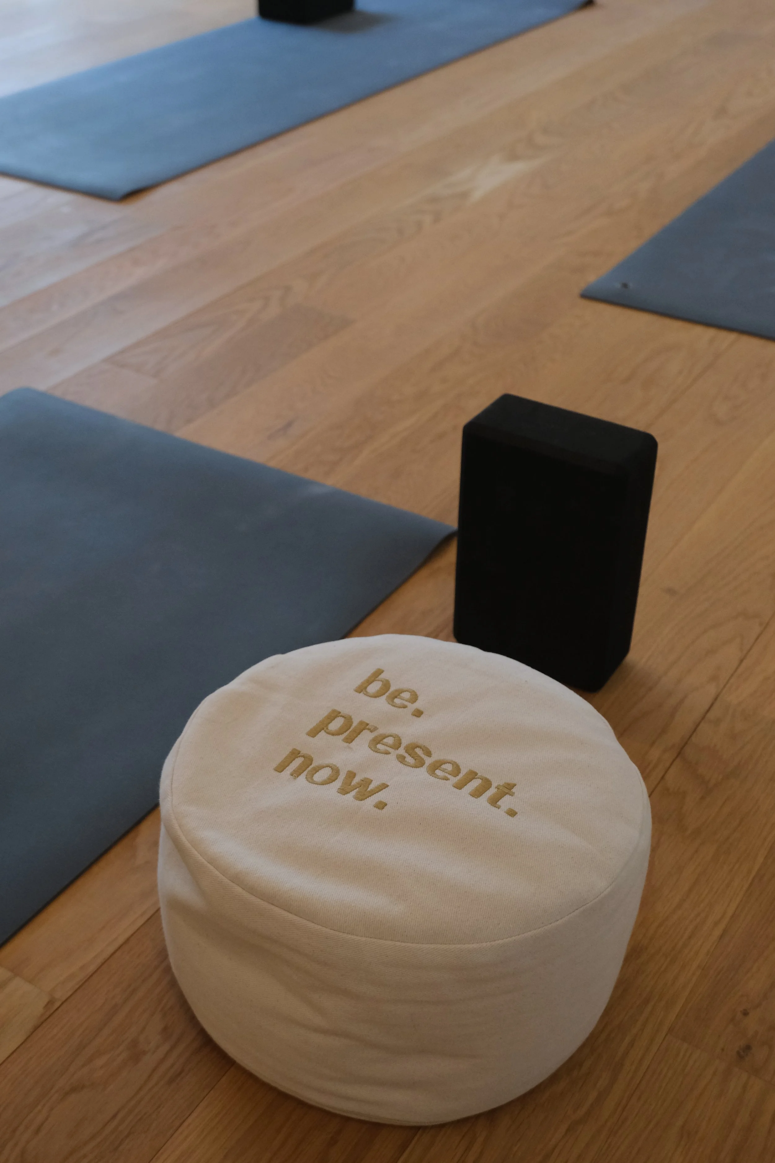 Ein rundes, weißes Meditationskissen mit goldener Aufschrift 'be. present. now.' auf einem Holzboden, umgeben von blauen Yogamatten und einem schwarzen Lautsprecher.