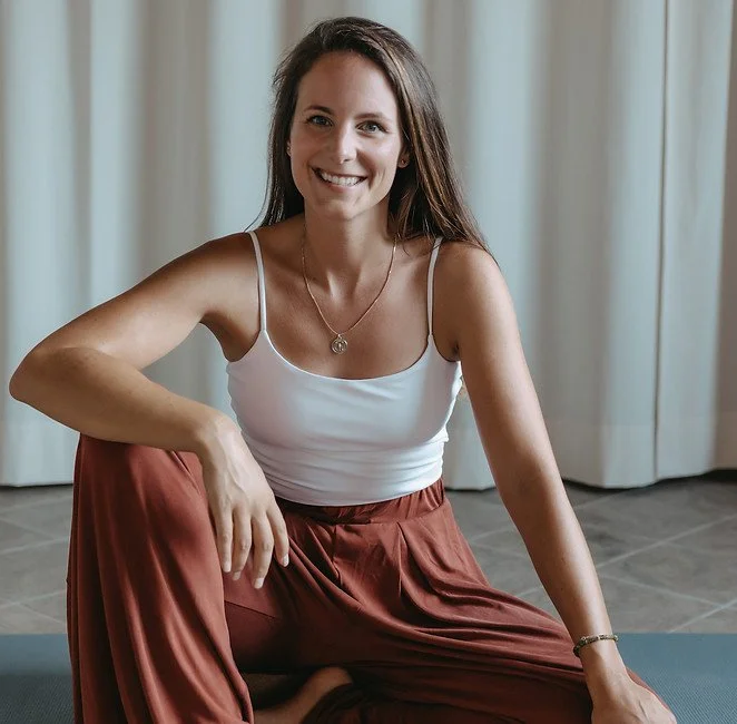 Eine lächelnde Frau sitzt auf einer Yogamatte in einem Raum mit hellen Vorhängen, trägt ein weißes Top und weite rote Hosen.