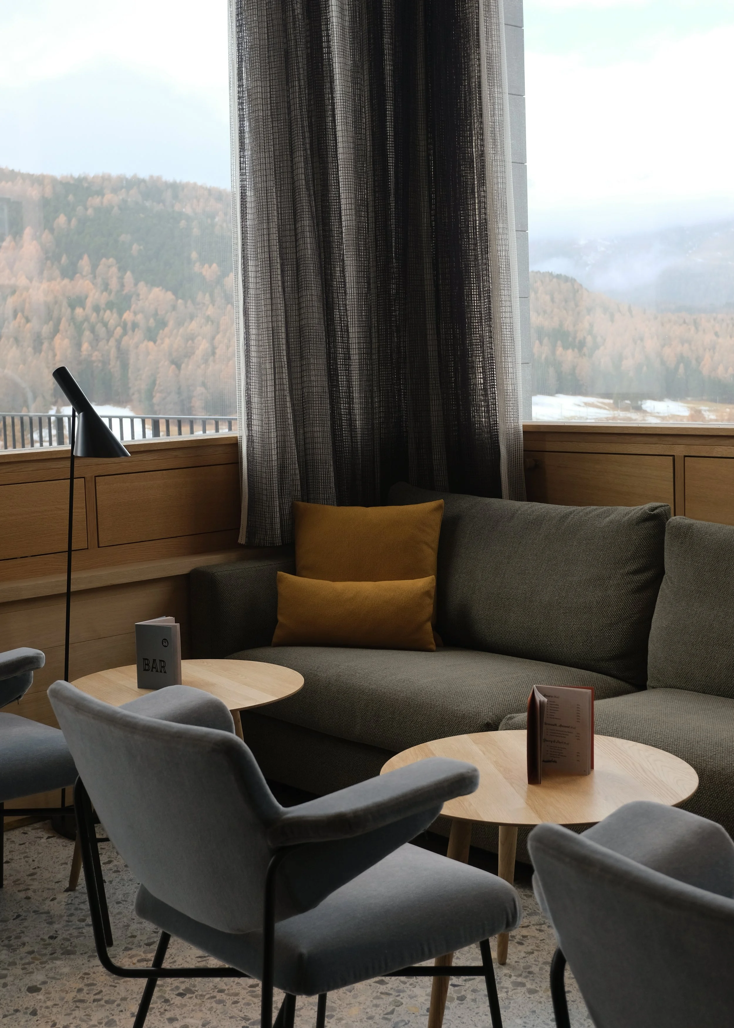 Innenansicht eines modernen Wohnzimmers mit grauem Sofa, dunklen Vorhängen, weißen Sesseln, Holztischen und Blick auf die Berglandschaft durch große Fenster
