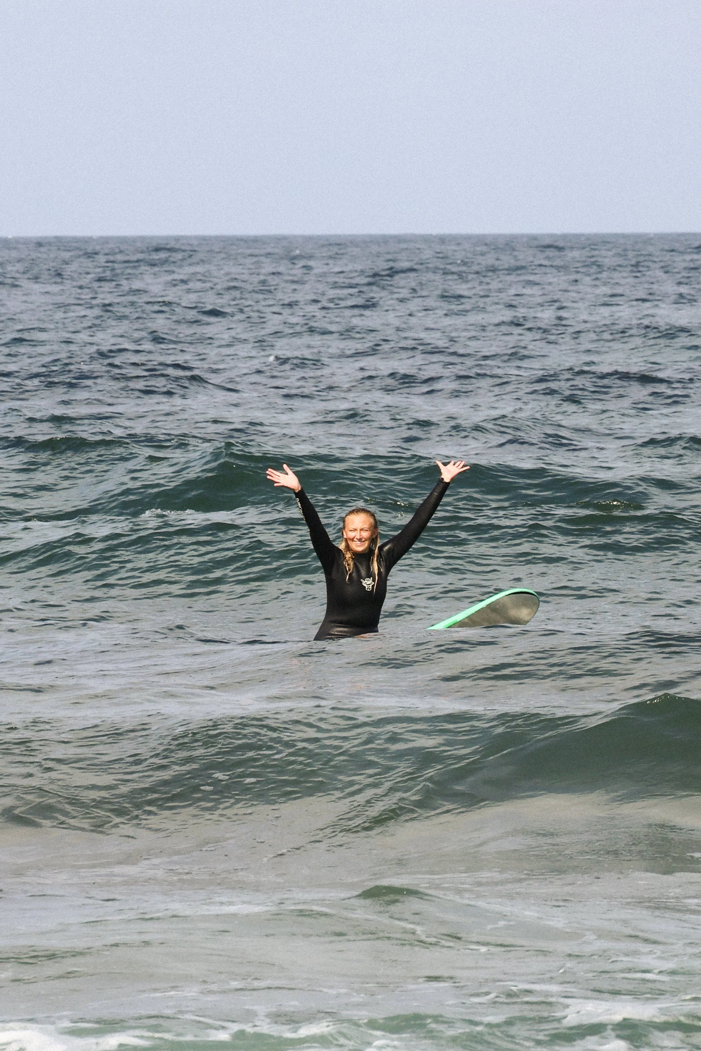 Frau im Meereswasser mit ausgestreckten Armen, lachend, Surfausrüstung, bei ruhigem Wasser. Horizont und Himmel im Hintergrund.
