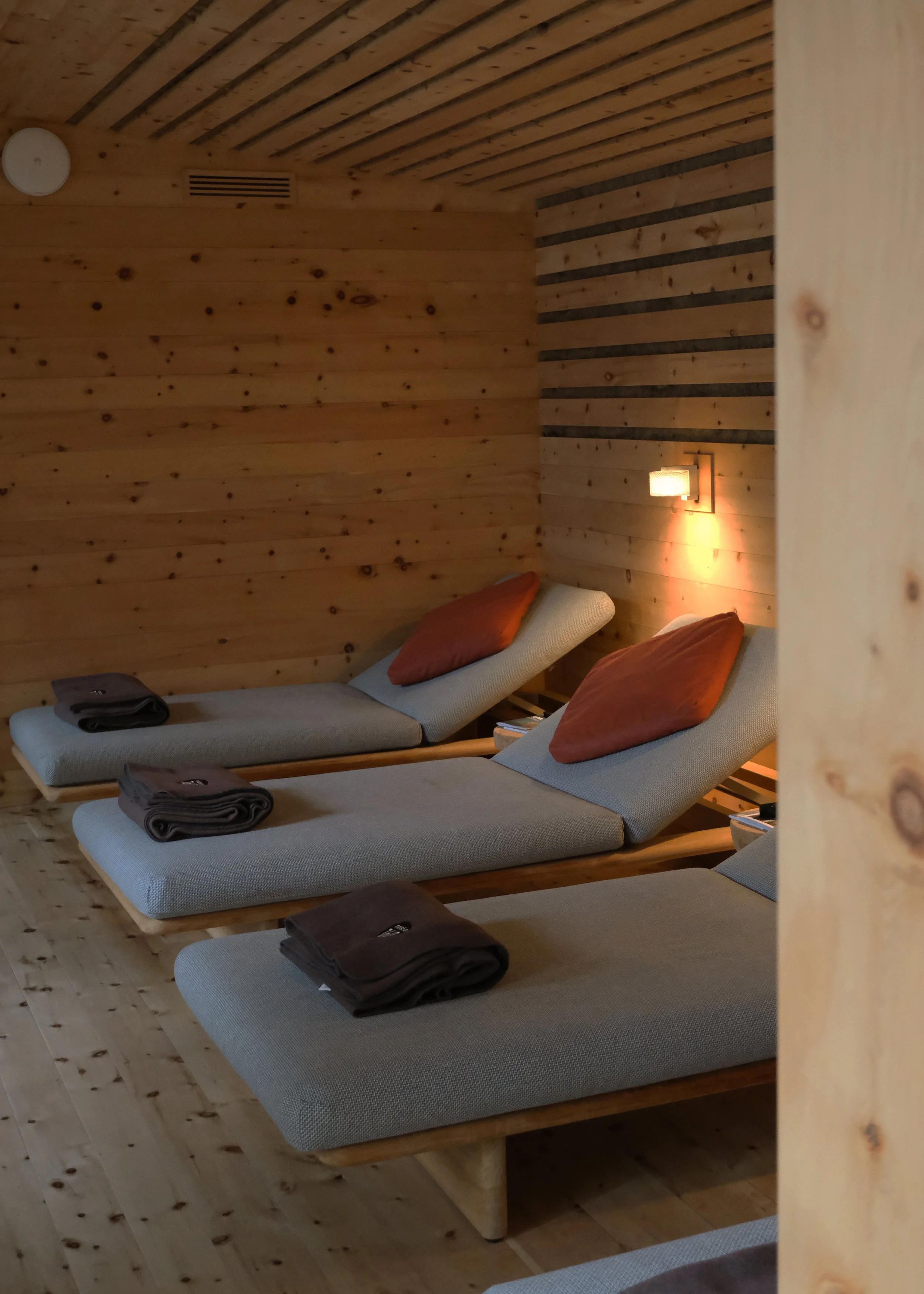 Drei Liegen mit grauer Polsterung und roten Kissen in einem Holzraum, jeweils mit Handtüchern darauf, an einer Wand mit Beleuchtung, Holzdecke und Holzfußboden.