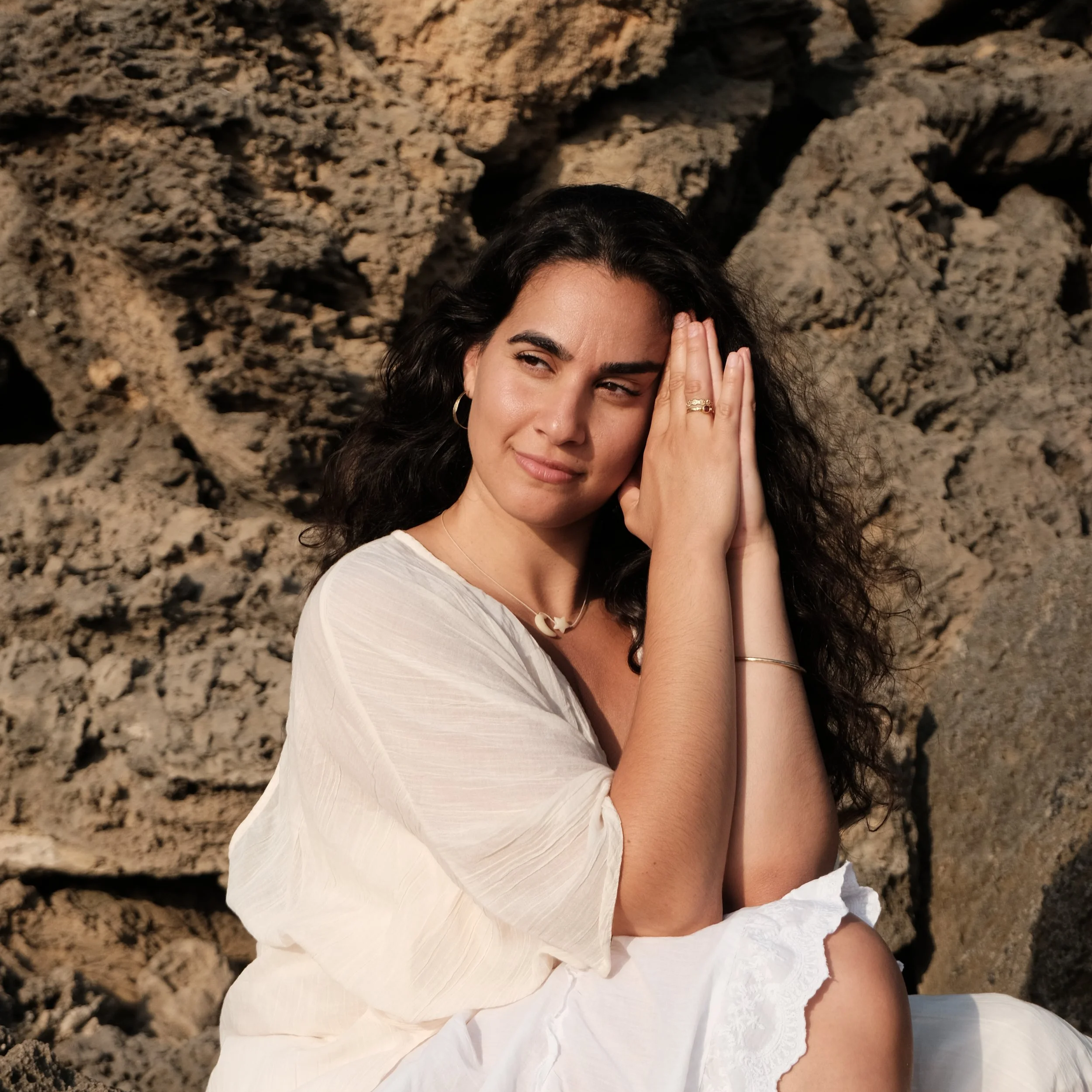 Junge Frau mit dunklen, lockigen Haaren, traut sich die Hand ans Gesicht, vor Felsen im Hintergrund.