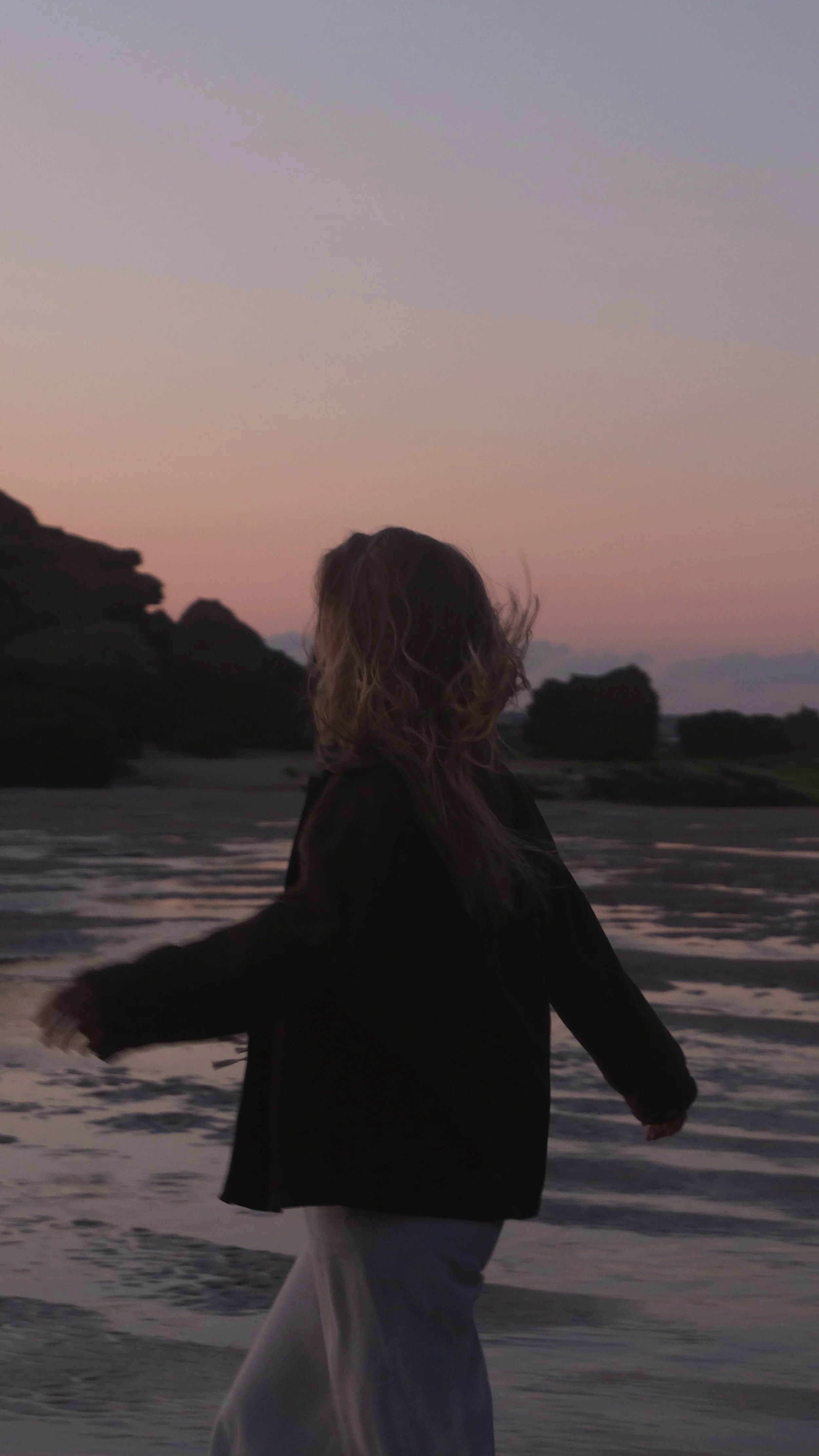 Eine Person mit lockigem Haar in einem schwarzen Mantel läuft am Strand bei Sonnenuntergang.