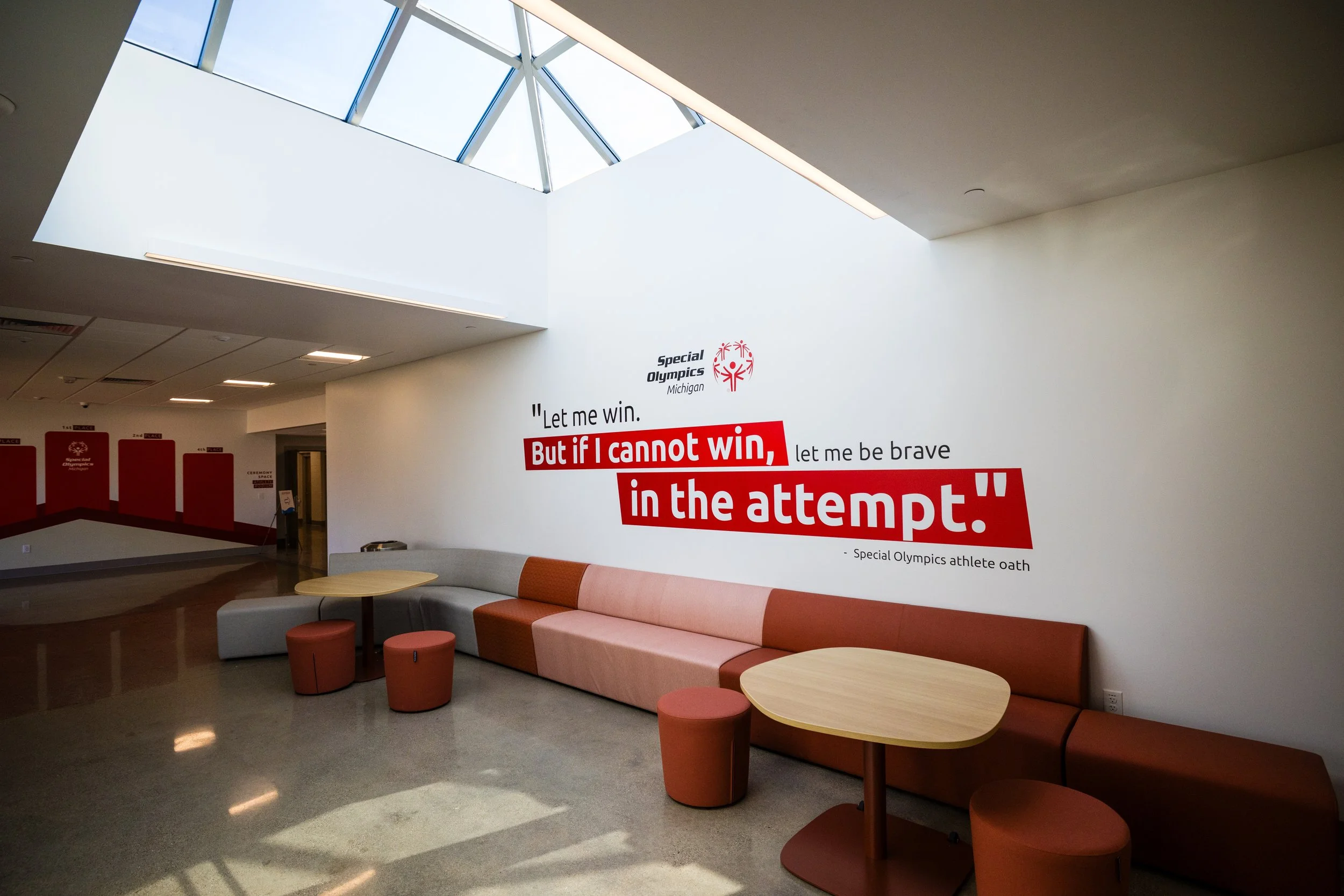 Lobby area of a facility with a large white wall, featuring a quote about winning and trying from a Special Olympics athlete, and the Special Olympics Michigan logo. There is a curved, multi-colored orange and beige sofa, round tables, and orange upholstered stools. A skylight provides natural light.