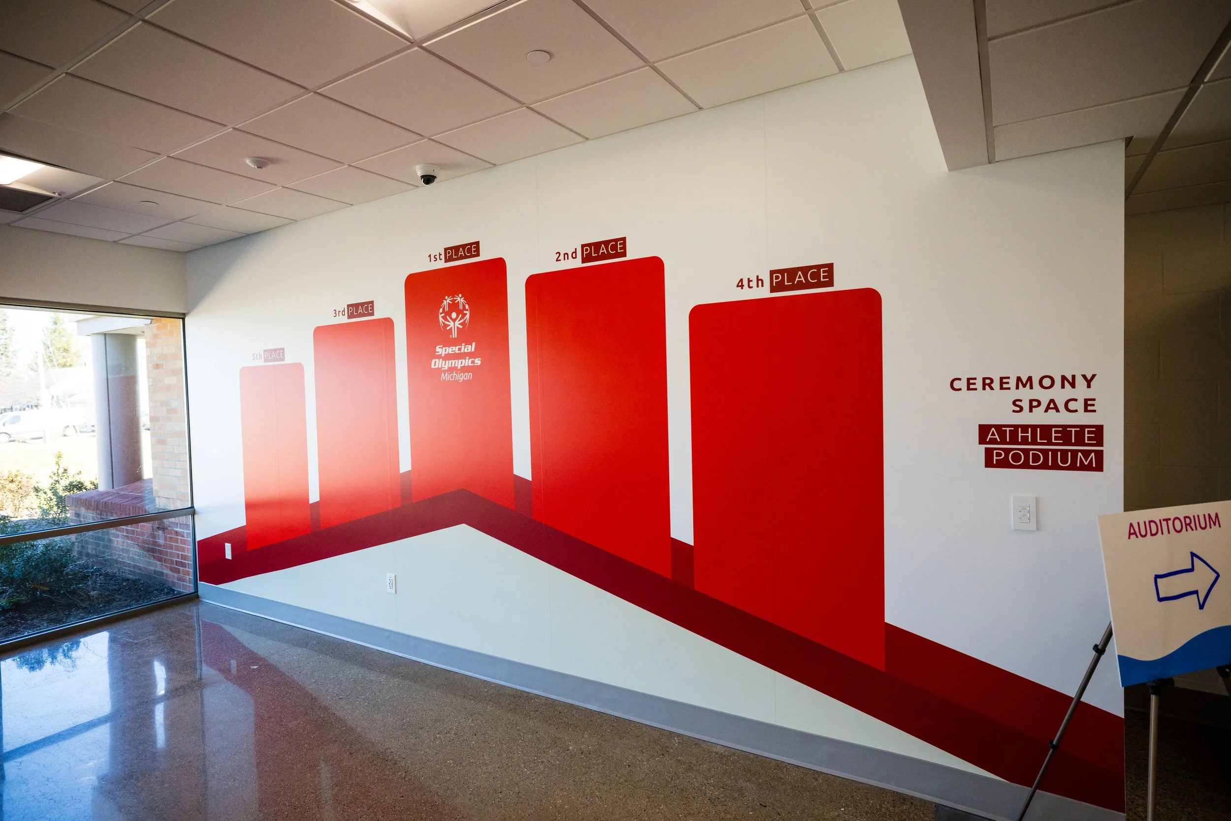 Wall with red and white graphic showing 1st to 5th place standings for Special Olympics Michigan, with a section labeled 'Ceremony Space Athlete Podium'; sign pointing to the auditorium.