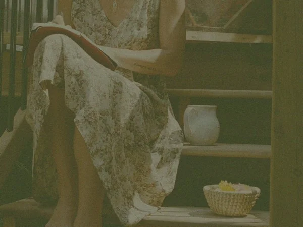 Woman sitting on wooden steps writing in journal with tea and small wicker basket beside her