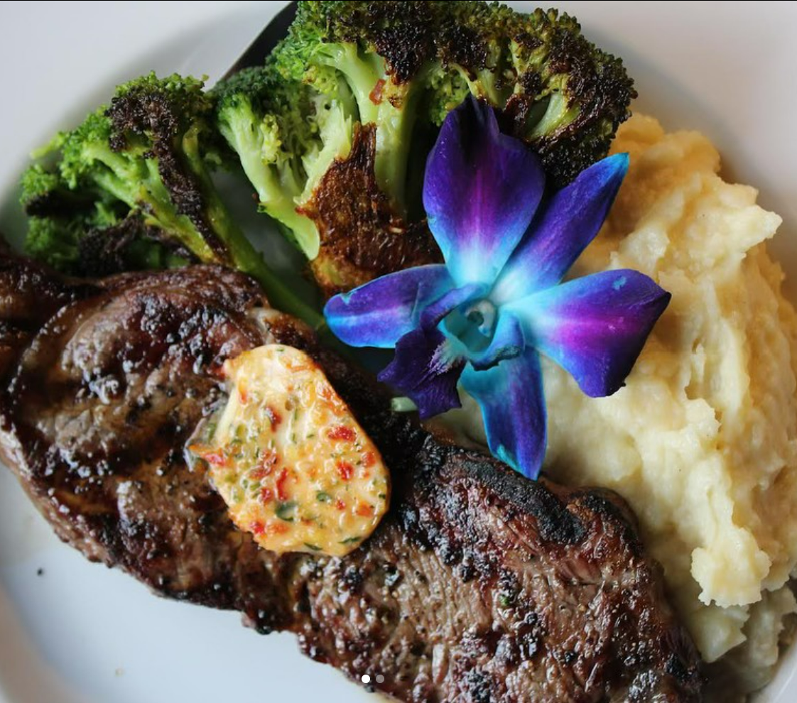A plate of grilled steak topped with herb butter, served with roasted broccoli, mashed potatoes, and a purple orchid flower for garnish.