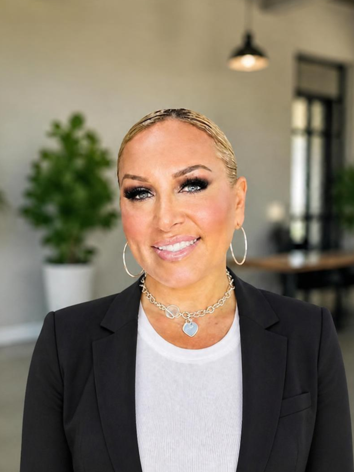 A woman with blonde hair, wearing a black blazer and jewelry, standing outdoors next to a tree and smiling.
