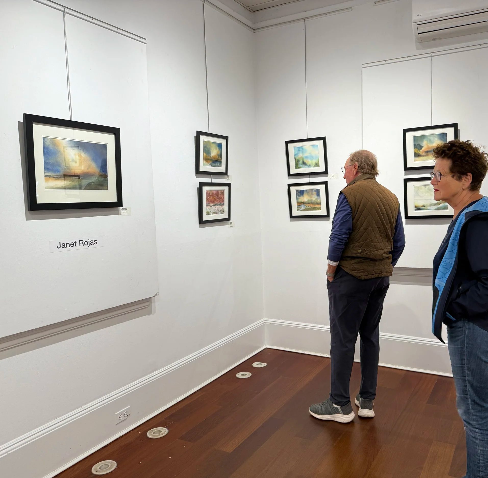 Two people viewing framed landscape paintings at an art gallery, with a sign that reads 'Janet Rojas' on the wall.