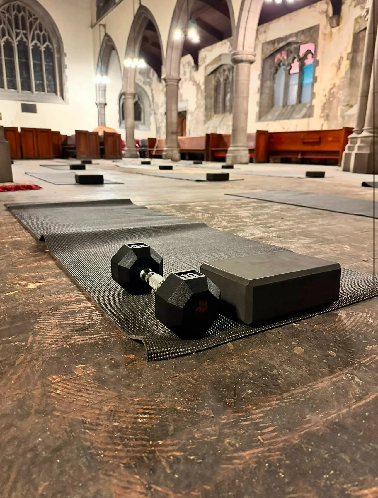 Yoga mat, dumbbells, and yoga blocks set up on the floor inside a church or historical building with stone pillars and stained glass windows.