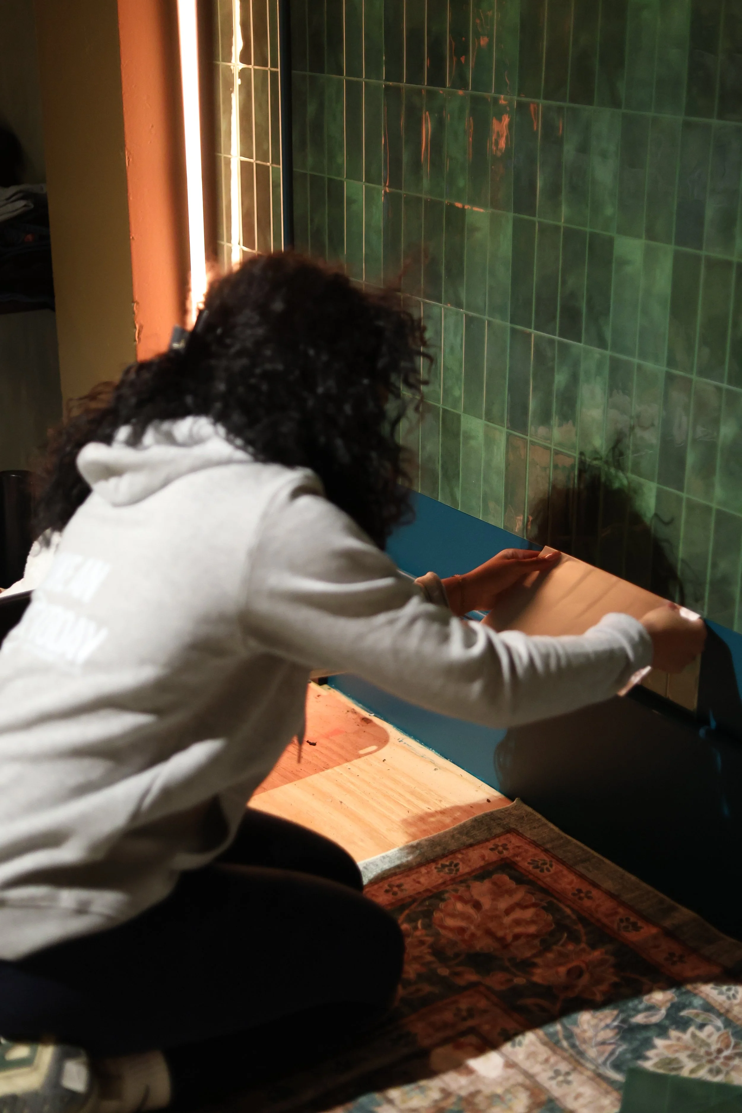Person kneeling on a patterned carpet, working on a large cardboard or paper on a table, with green tiled wall in the background.