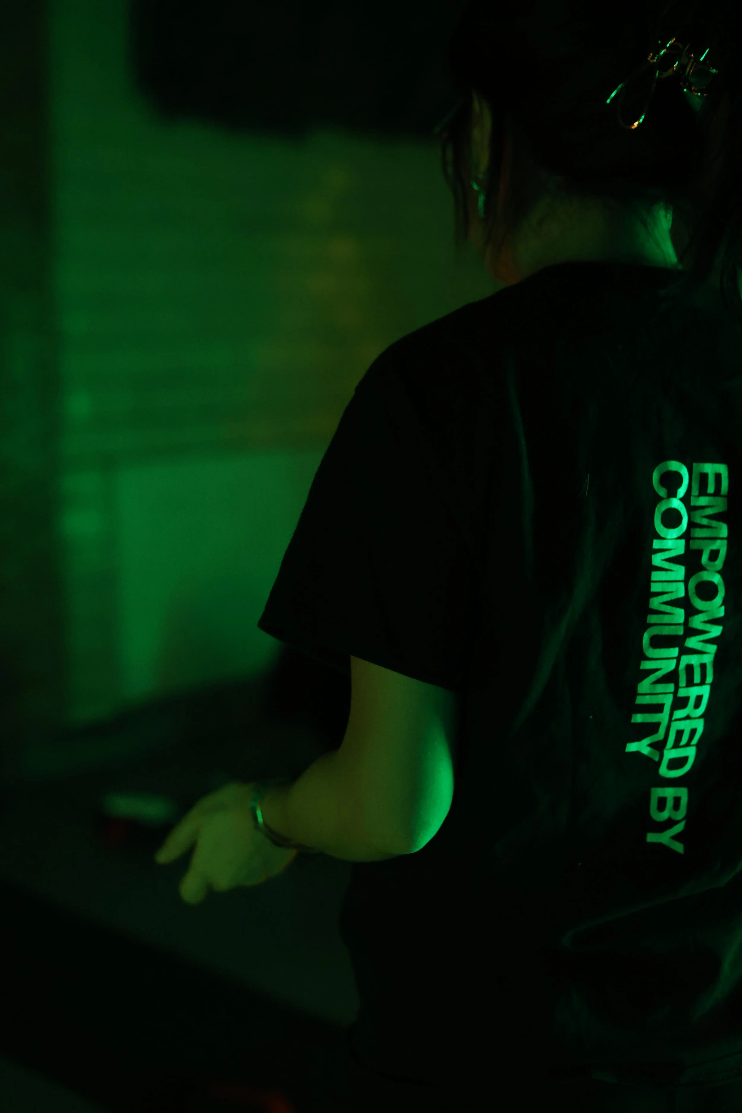 Person in a black shirt with green reflective text 'EMPLOYED BY COMMUNITY' on the back, wearing gloves, in a dark green-lit environment, facing away from the camera.