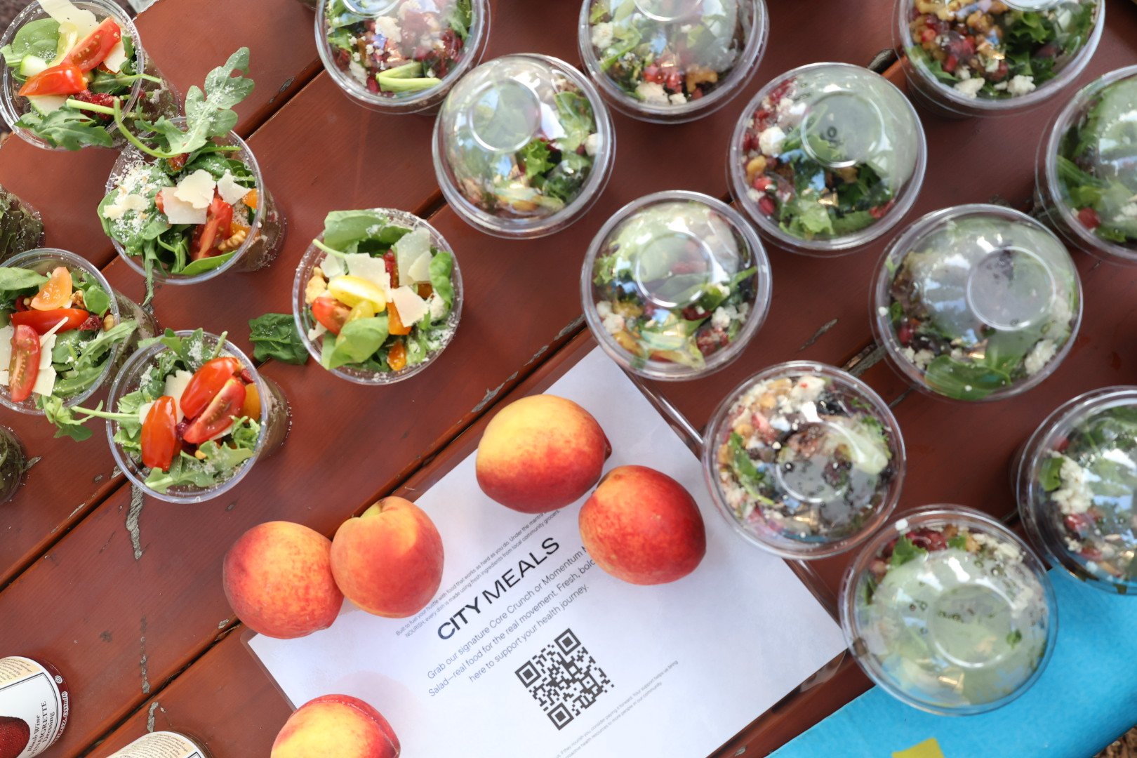 A table with multiple plastic cups filled with fresh salad containing lettuce, cherry tomatoes, cheese, and other vegetables, along with three peaches, a paper with the title 'City Meals' and a QR code, and a partial view of a menu and a can.