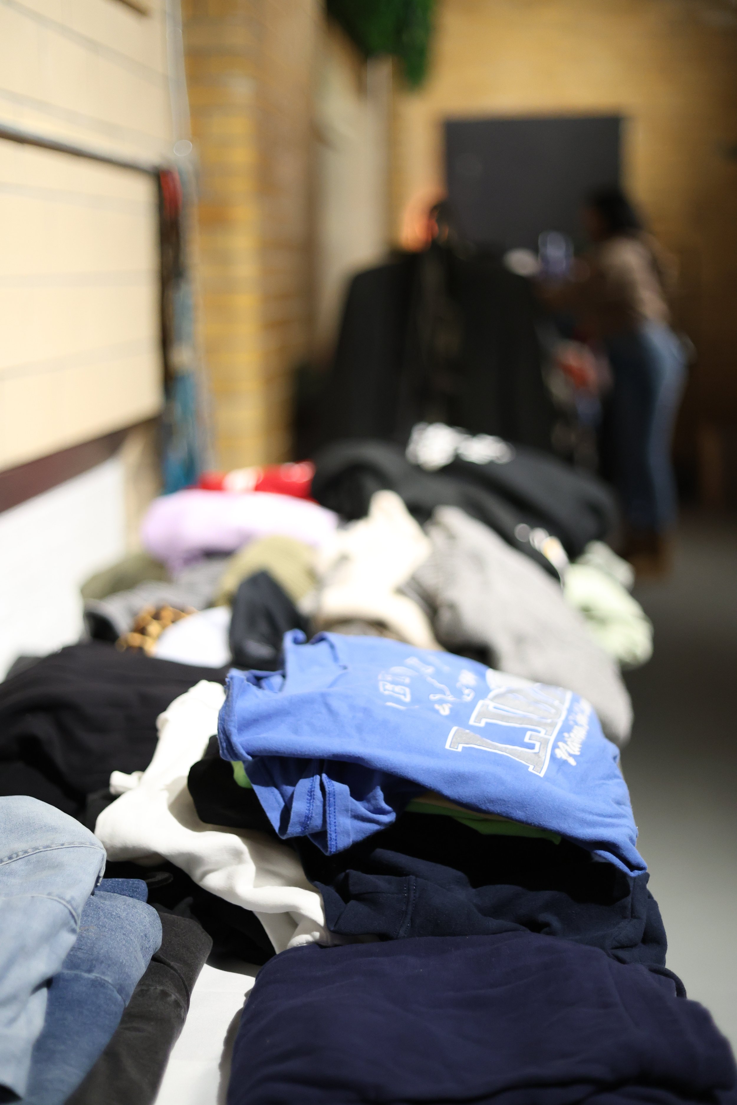 A pile of various folded and unfolded clothes on a table in an indoor setting, with two people blurred in the background.