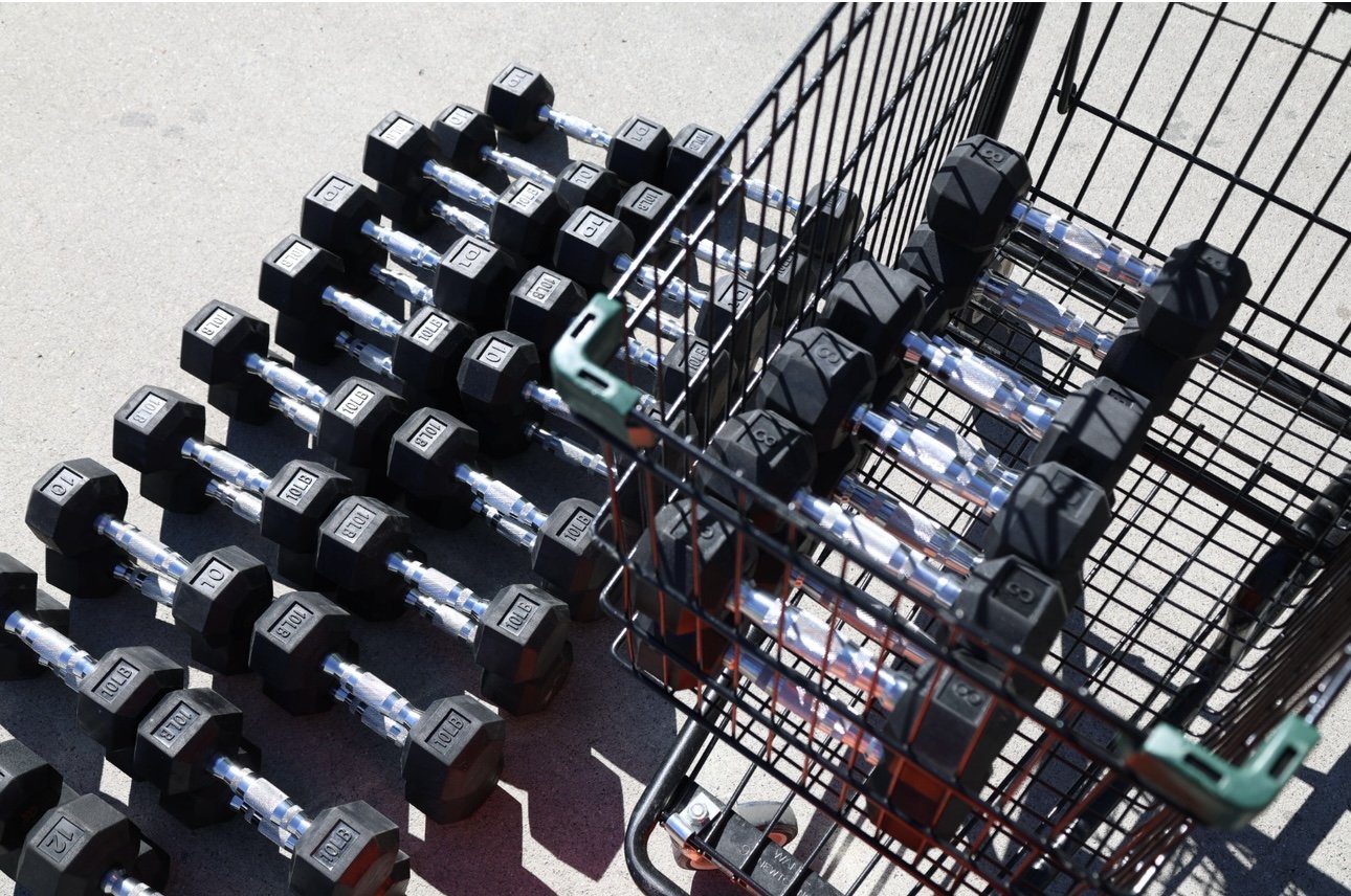 A shopping cart containing multiple dumbbells, with more dumbbells lined up on the ground nearby.