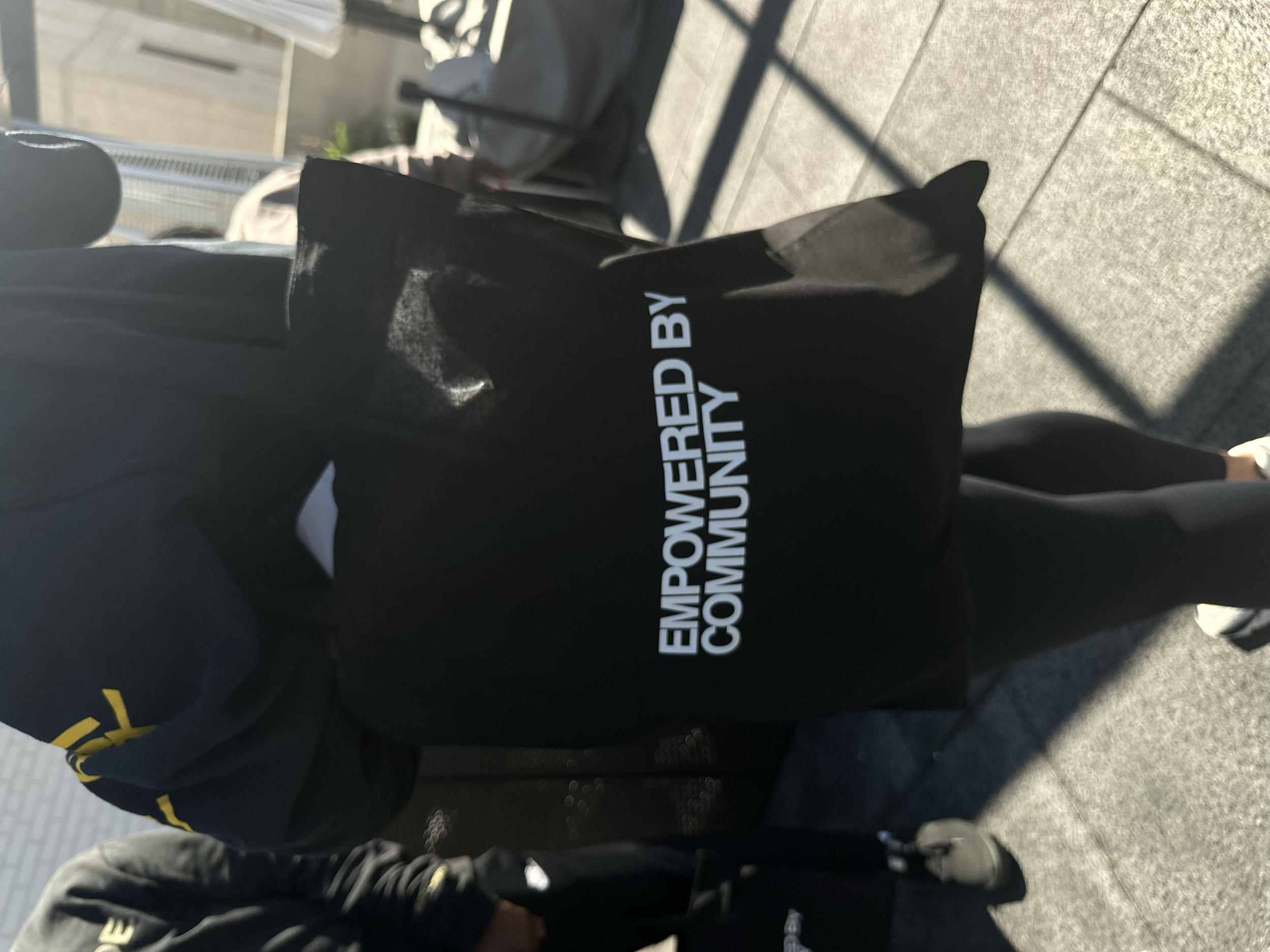 Person holding a black tote bag with white text that reads 'Empowered by Community' while standing outside on a sidewalk.