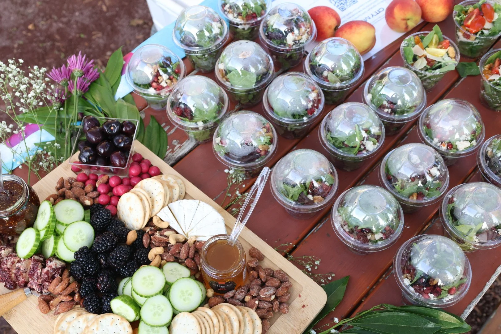 A picnic table with a variety of fresh fruit, crackers, and honey, alongside several bowls of mixed salad in clear plastic cups.