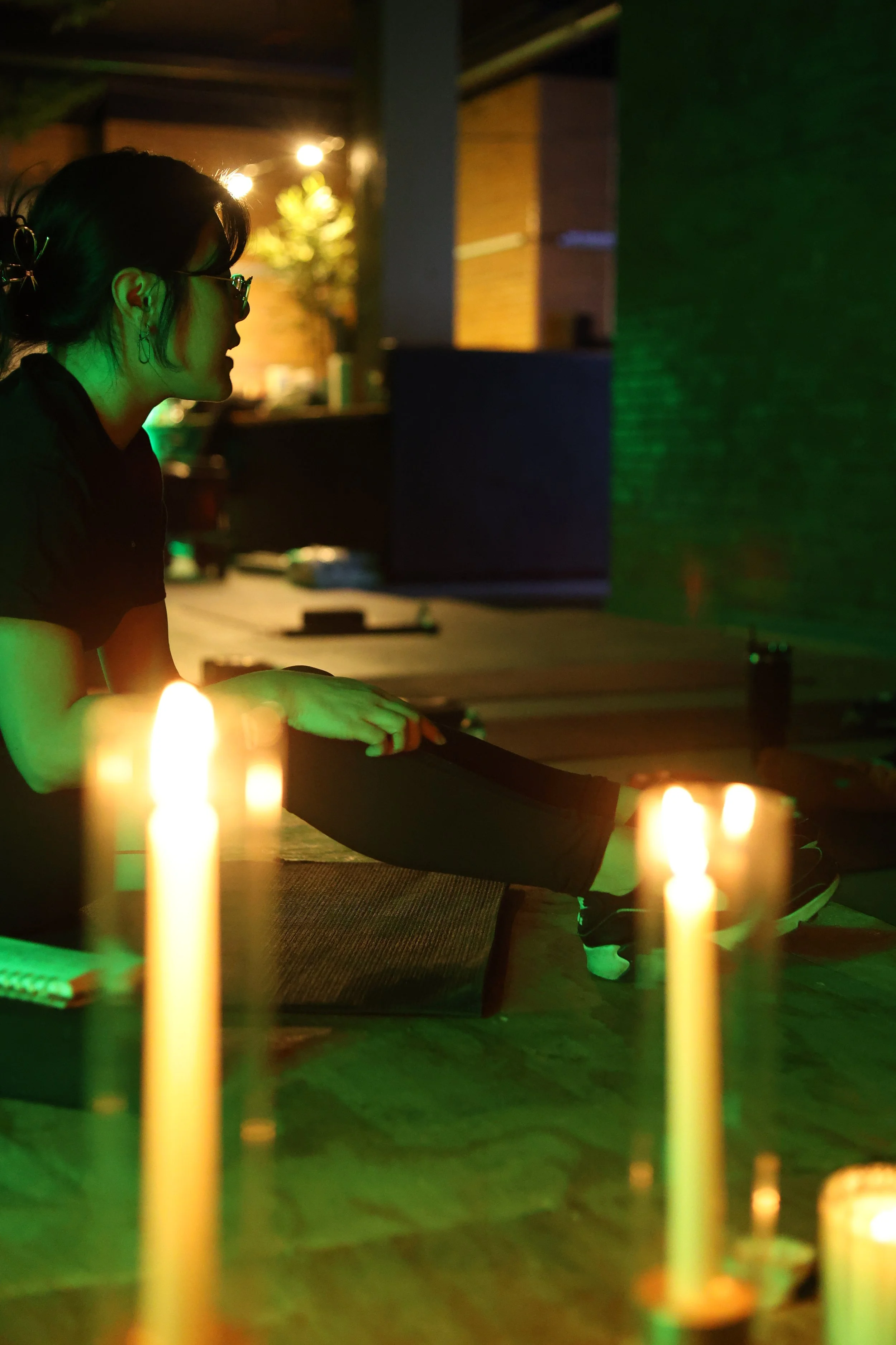 A woman practicing yoga or meditation at night with candles in front of her, illuminated by green and warm lighting.