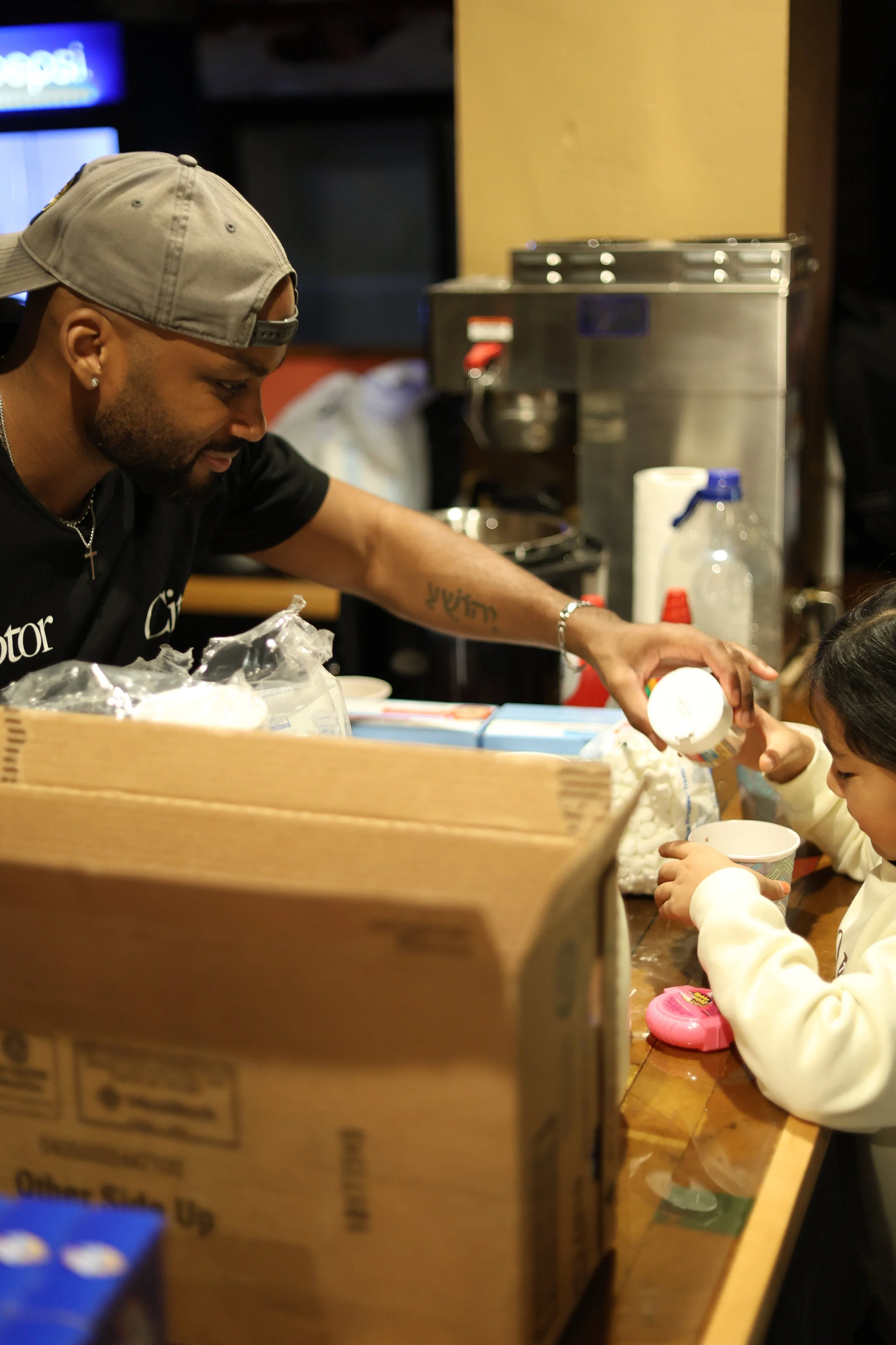 A man in a gray baseball cap and black shirt is pouring a white powder into a child's cup at a wooden table in a casual indoor setting.