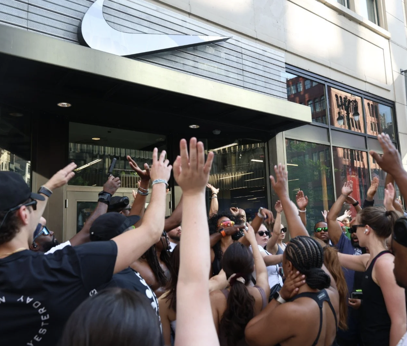 A crowd of diverse people gathered outside a building with their hands raised, participating in a group activity or protest.
