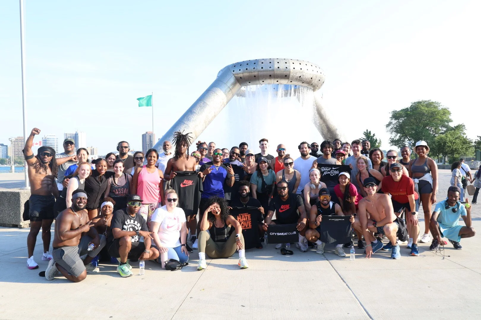 Group of diverse people posing in front of a large water fountain on a city street, some making peace signs, smiling, and wearing athletic clothing and sunglasses.