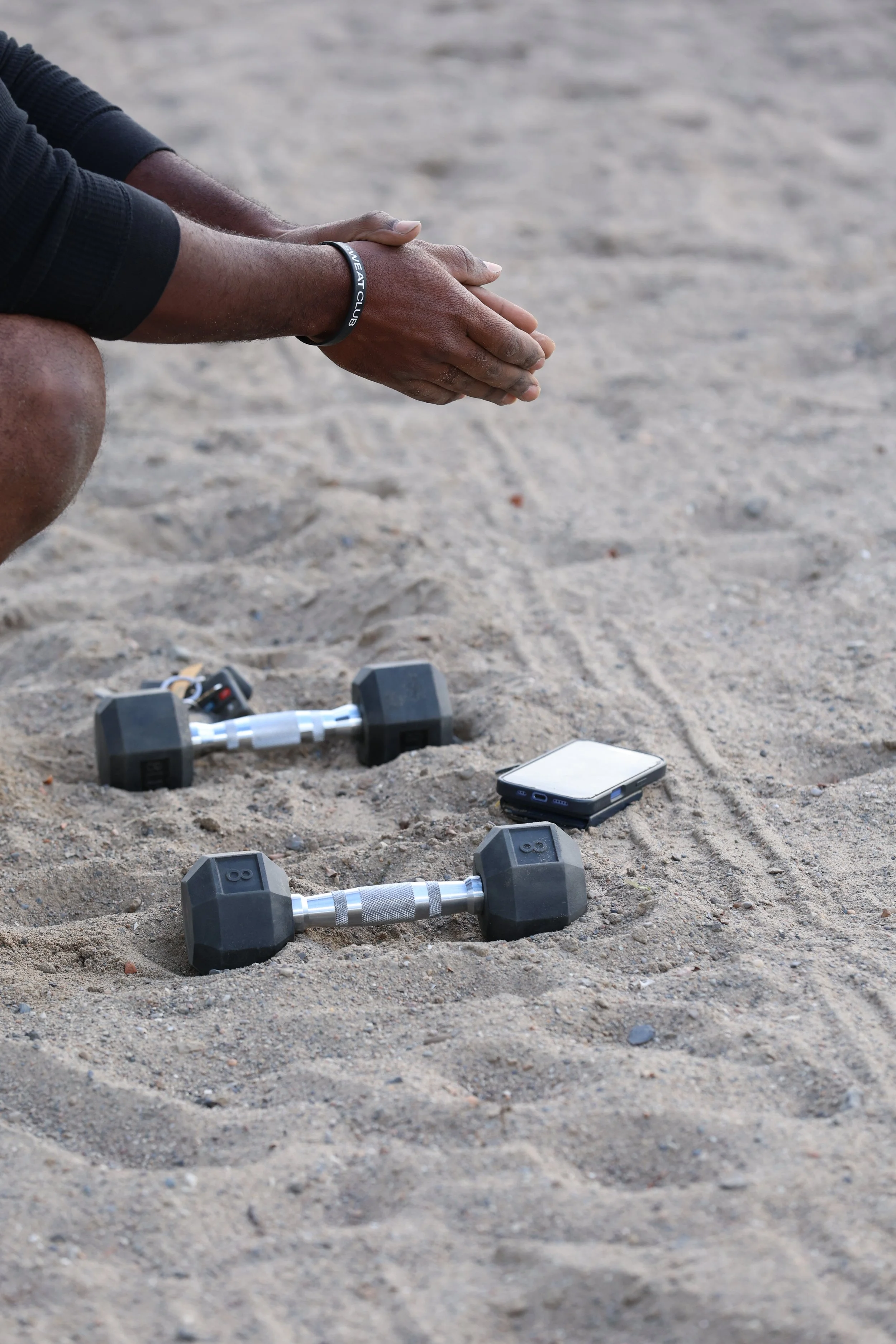 Person wearing a fitness club bracelet kneeling on sandy ground with two dumbbells and a smartphone placed on the sand.