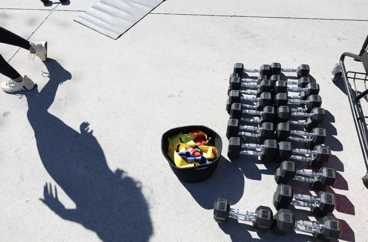 Dumbbells and kettlebells arranged on the ground outside, with a person's shadow and a shopping cart nearby.